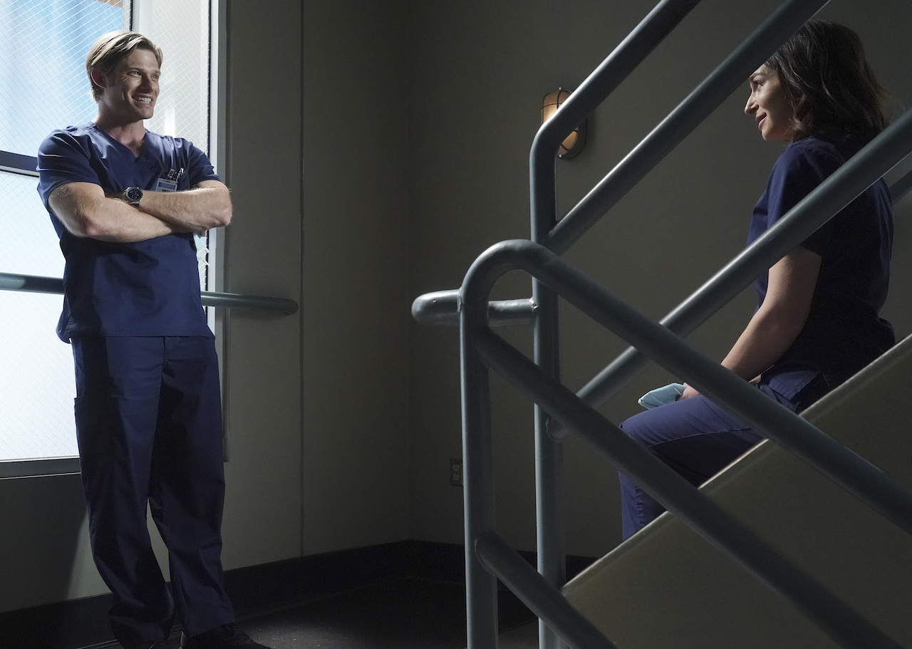 Chris Carmack as Link stands while talking to Caterina Scorsone as Amelia as she sits on stairs in 'Grey's Anatomy'