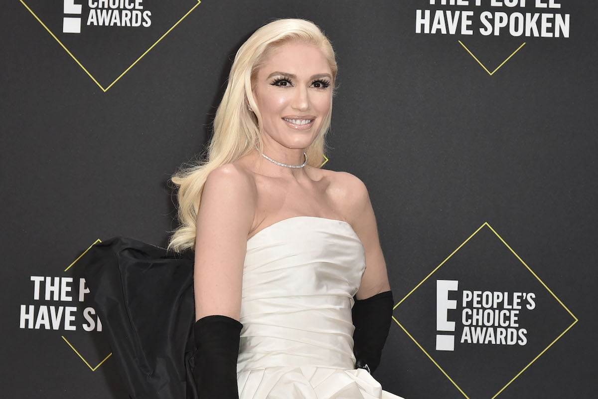Gwen Stefani smiles and poses in a white dress and black gloves at an event.