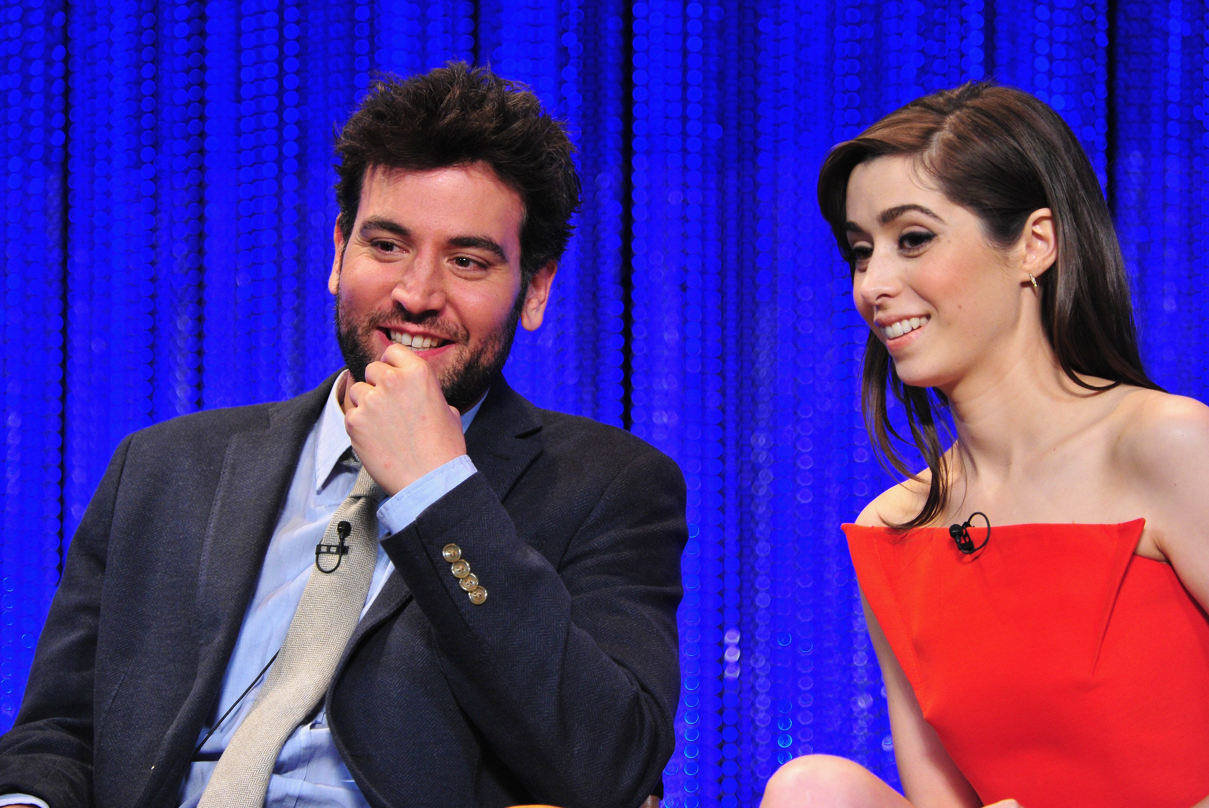 'How I Met Your Mother' stars Josh Radnor and Cristin Milioti sit next to one another during the panel. Radnor wears a dark blue suit over a light blue button-up shirt and light gray tie. Milioti wears a strapless red dress.