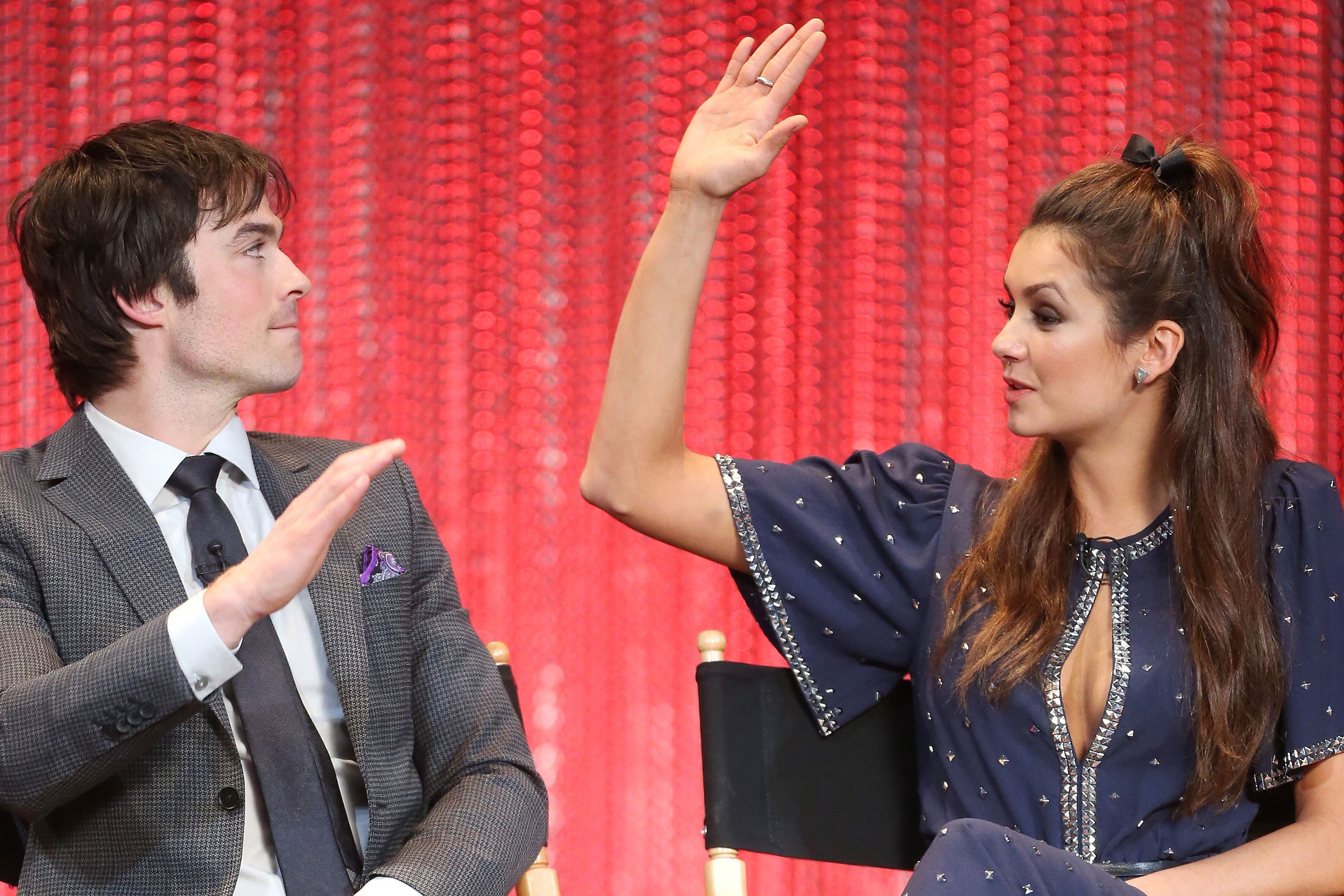 'The Vampire Diaries' stars Ian Somerhalder and Nina Dobrev raise their hands to high five. Somerhalder wears a gray suit over a white button-up shirt and black tie. Dobrev wears a blue dress with silver studs.