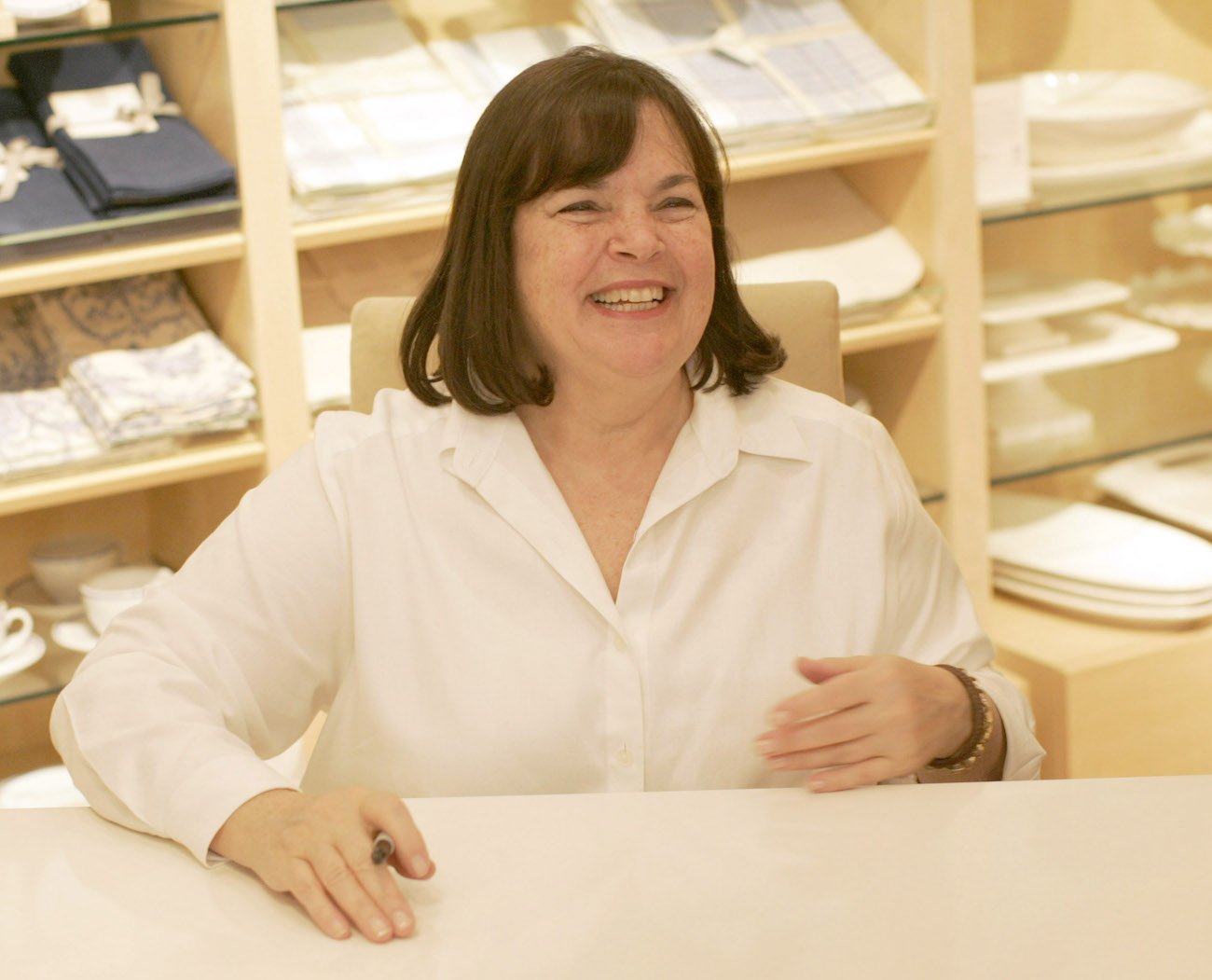 Ina Garten smiles wearing a white button-down shirt while sitting at a table