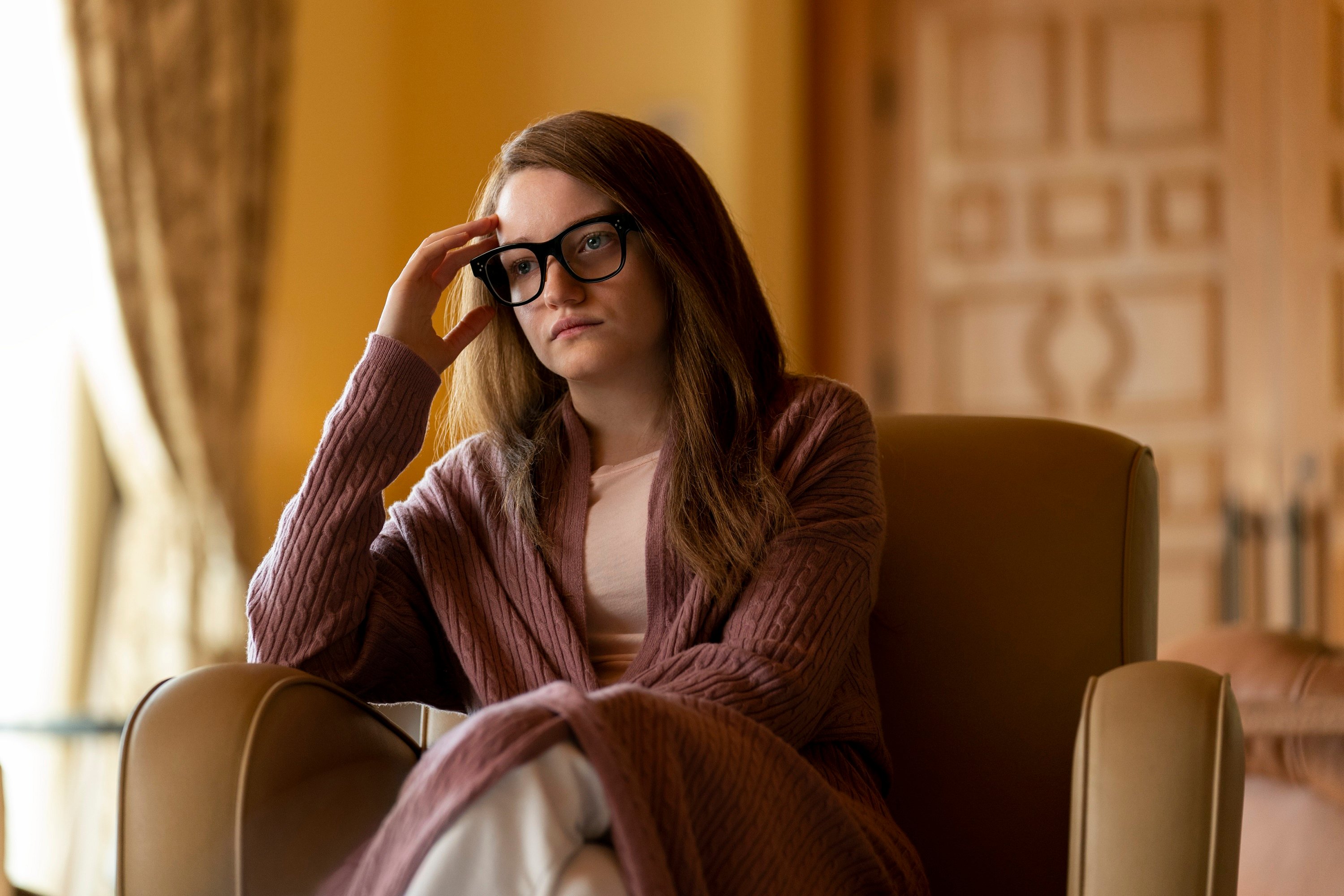 'Inventing Anna' cast member Julia Garner sits in a chair holding her glasses while looking upset as Anna Sorokin (Delvey)