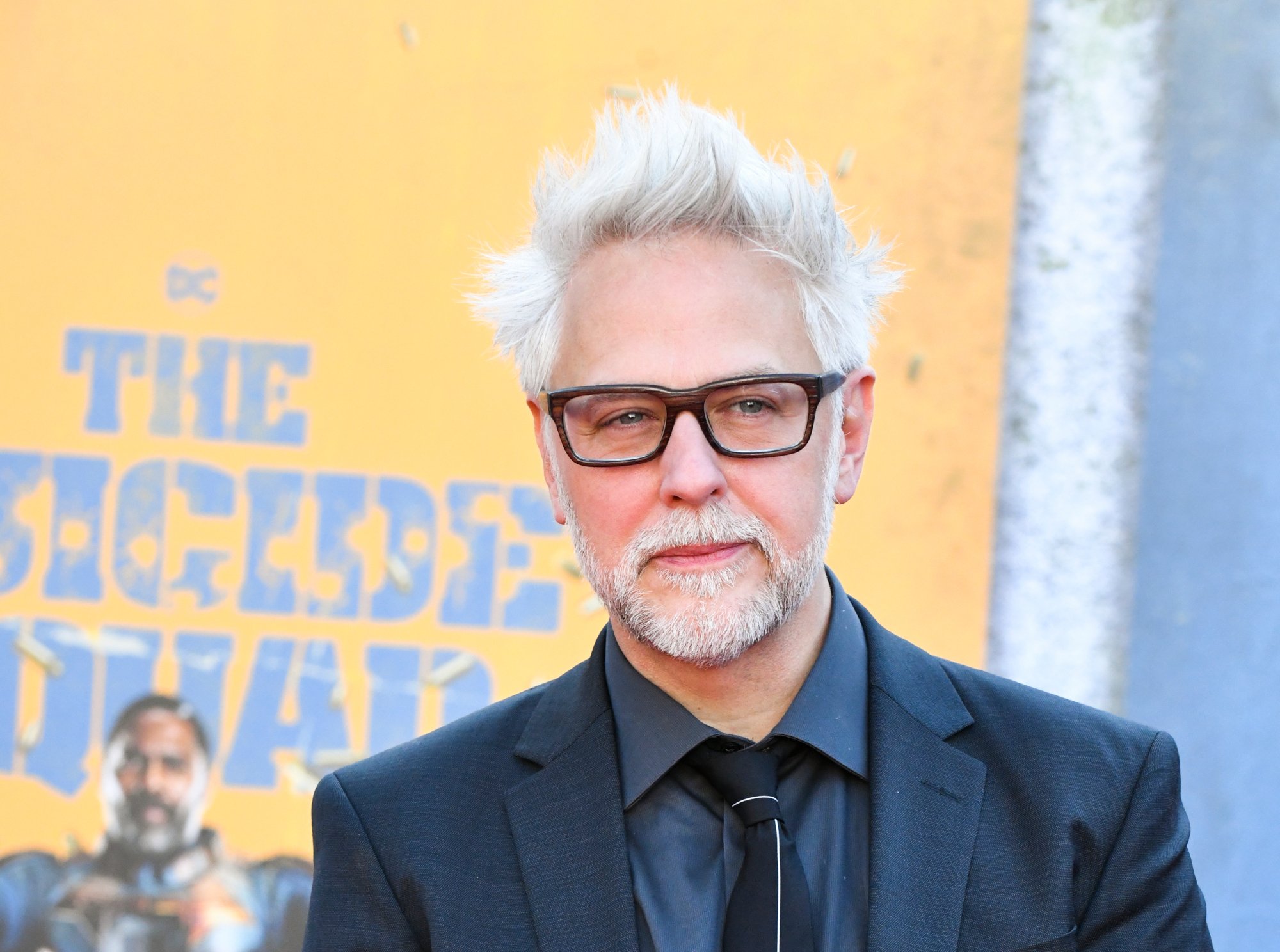 'The Suicide Squad' and 'Peacemaker' writer and director James Gunn. He's wearing a suit and glasses and standing in front of a colorful wall that says 'The Suicide Squad' on it.