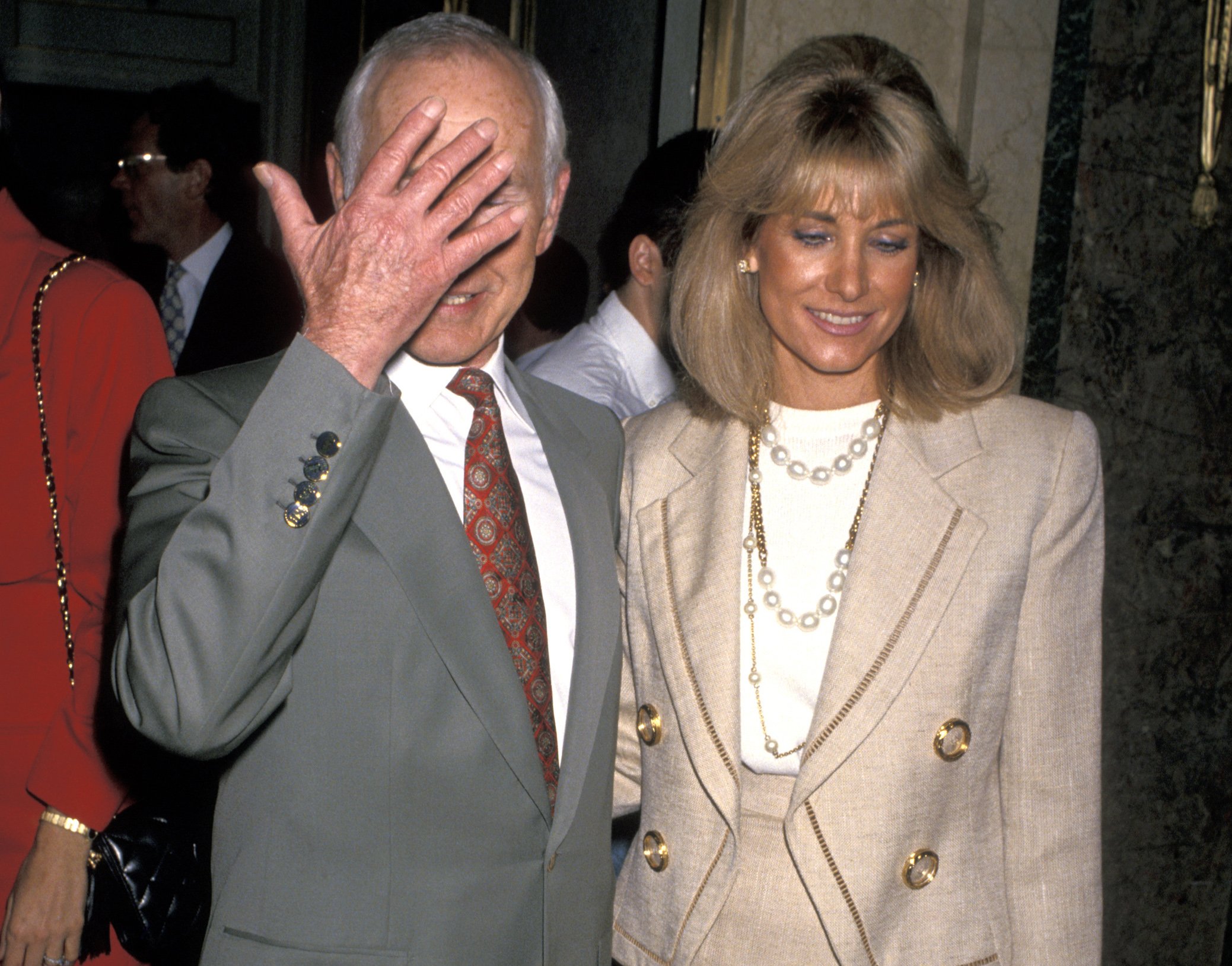 Johnny Carson in a gray suit holding a hand in front of his face while walking with wife, Alexis, in 1993