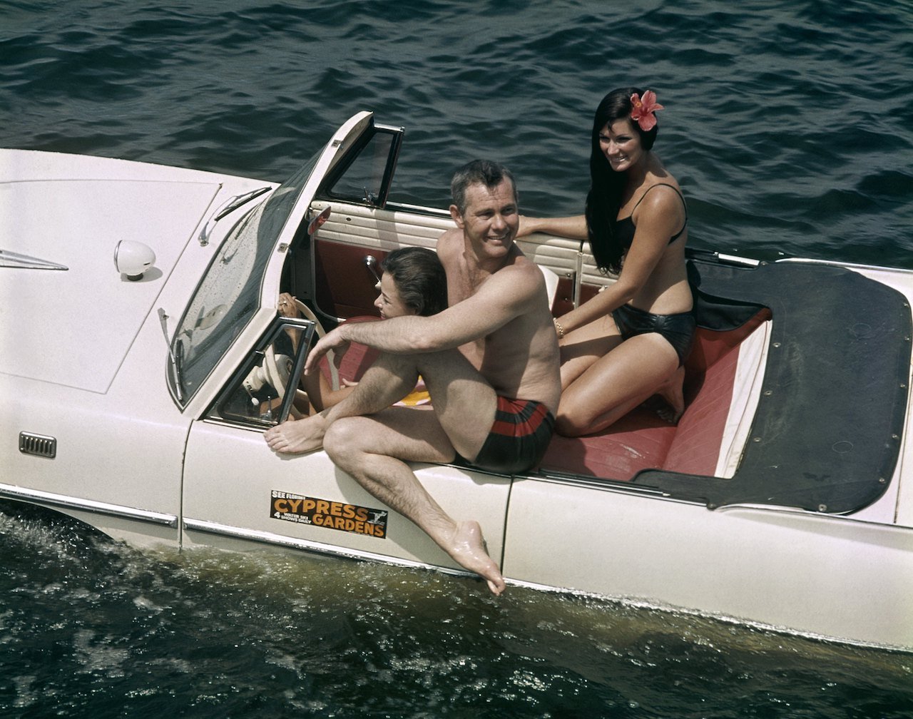 Johnny Carson sits in a car in the water with bikini-clad water skiers