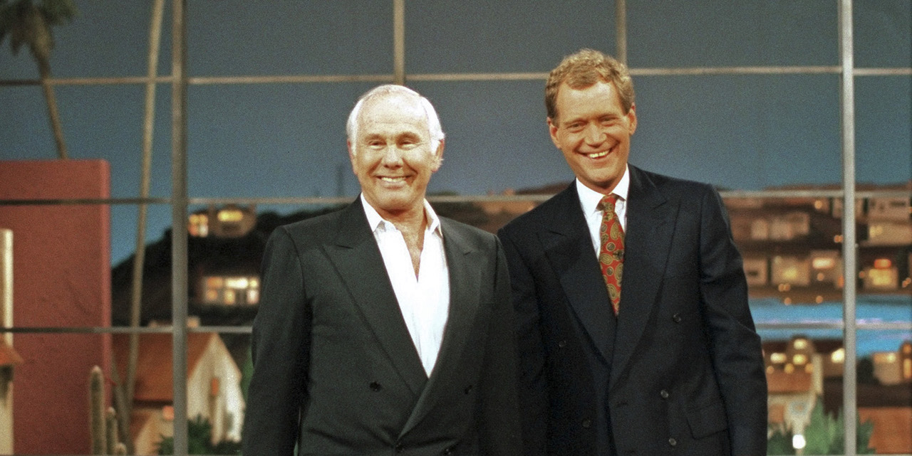 Johnny Carson in a black and white suit, standing next to David Letterman in a black suit with a red, patterned tie