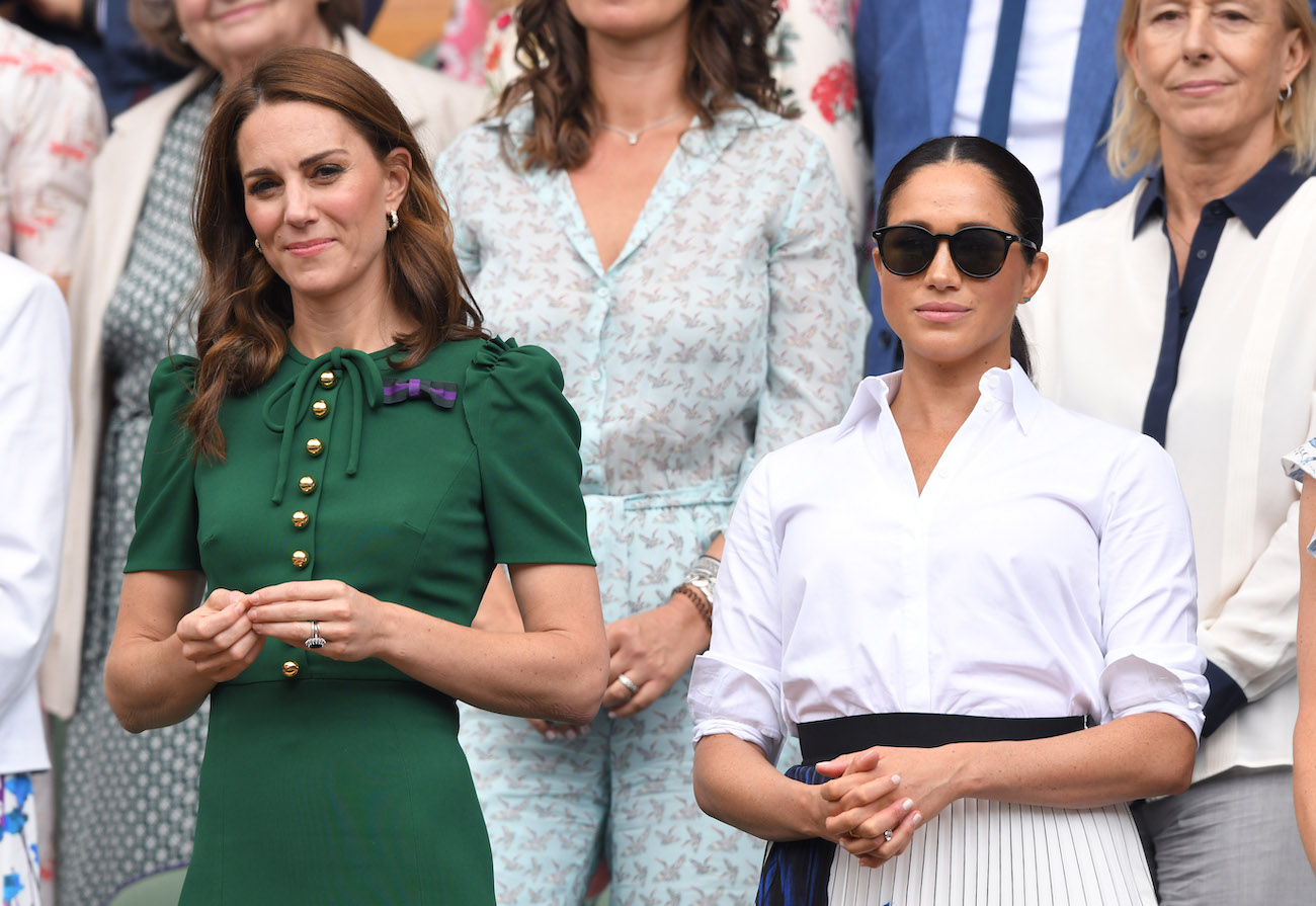 Kate Middleton wearing a green dress and standing next to Meghan Markle, who is wearing a white outfit and dark sunglasses