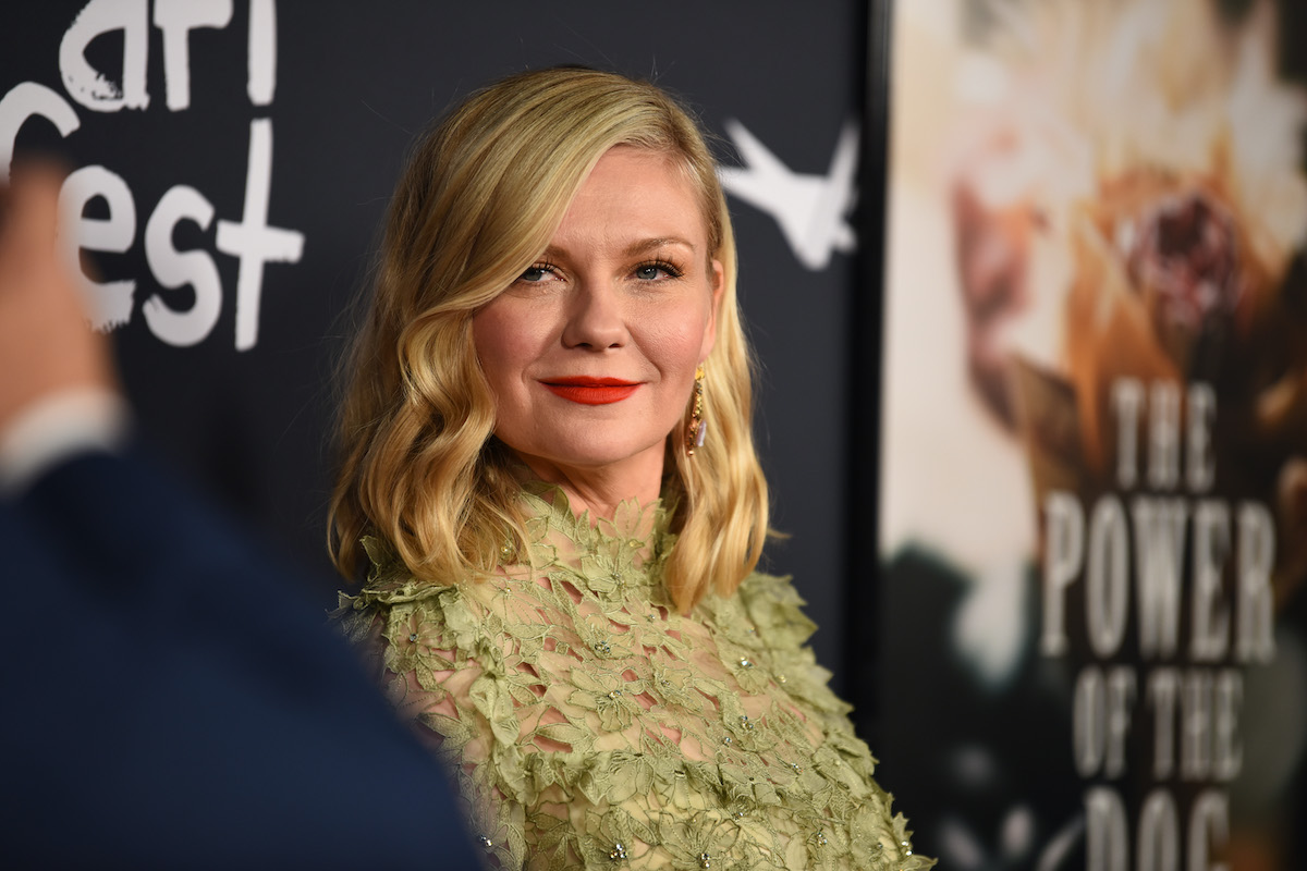 Kirsten Dunst wears a dress and poses with ‘The Power of the Dog’ logo in the background