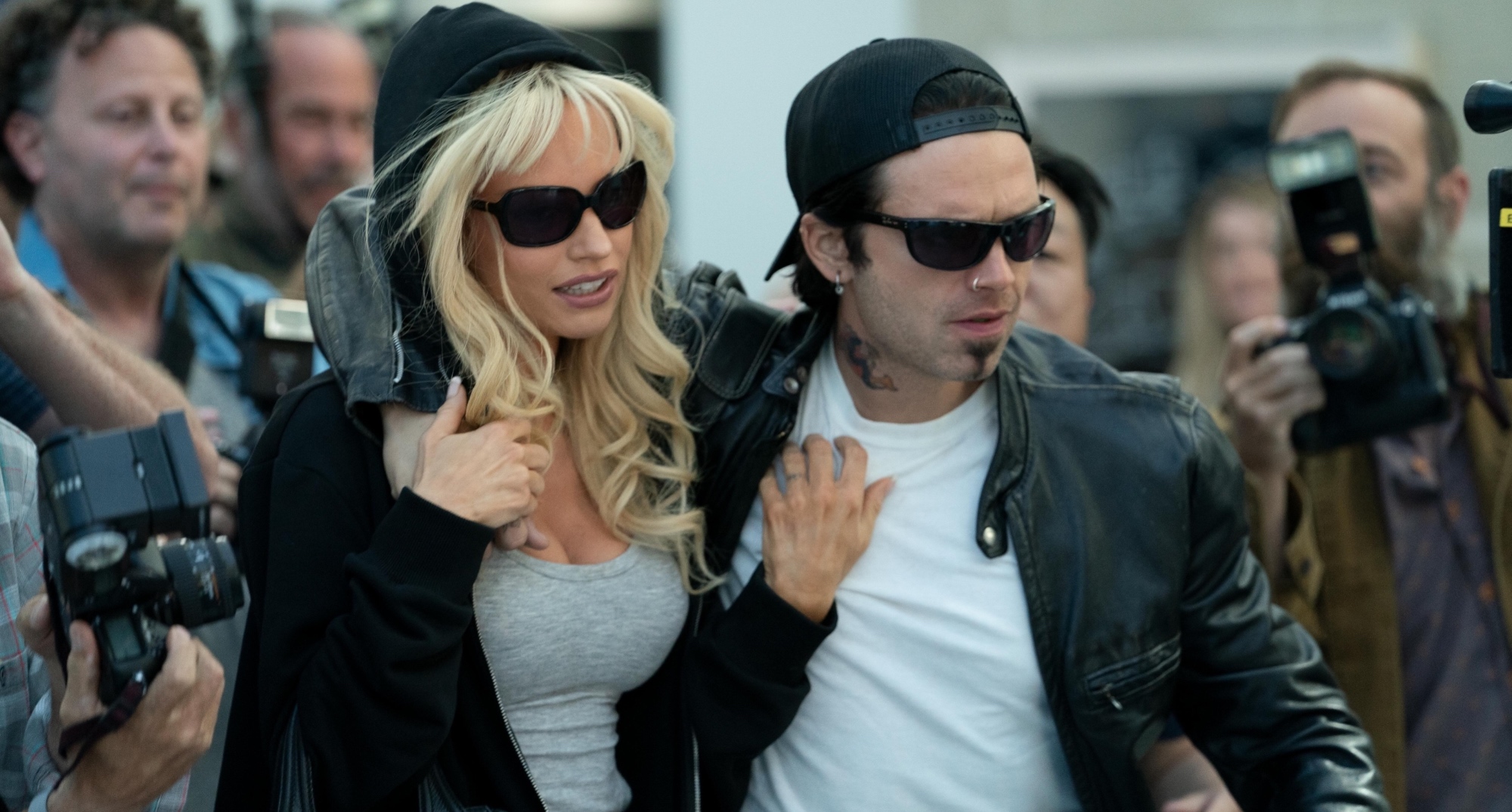 Lily James and Sebastian Stan in 'Pam & Tommy' series wearing sunglasses surrounded by paparazzi.
