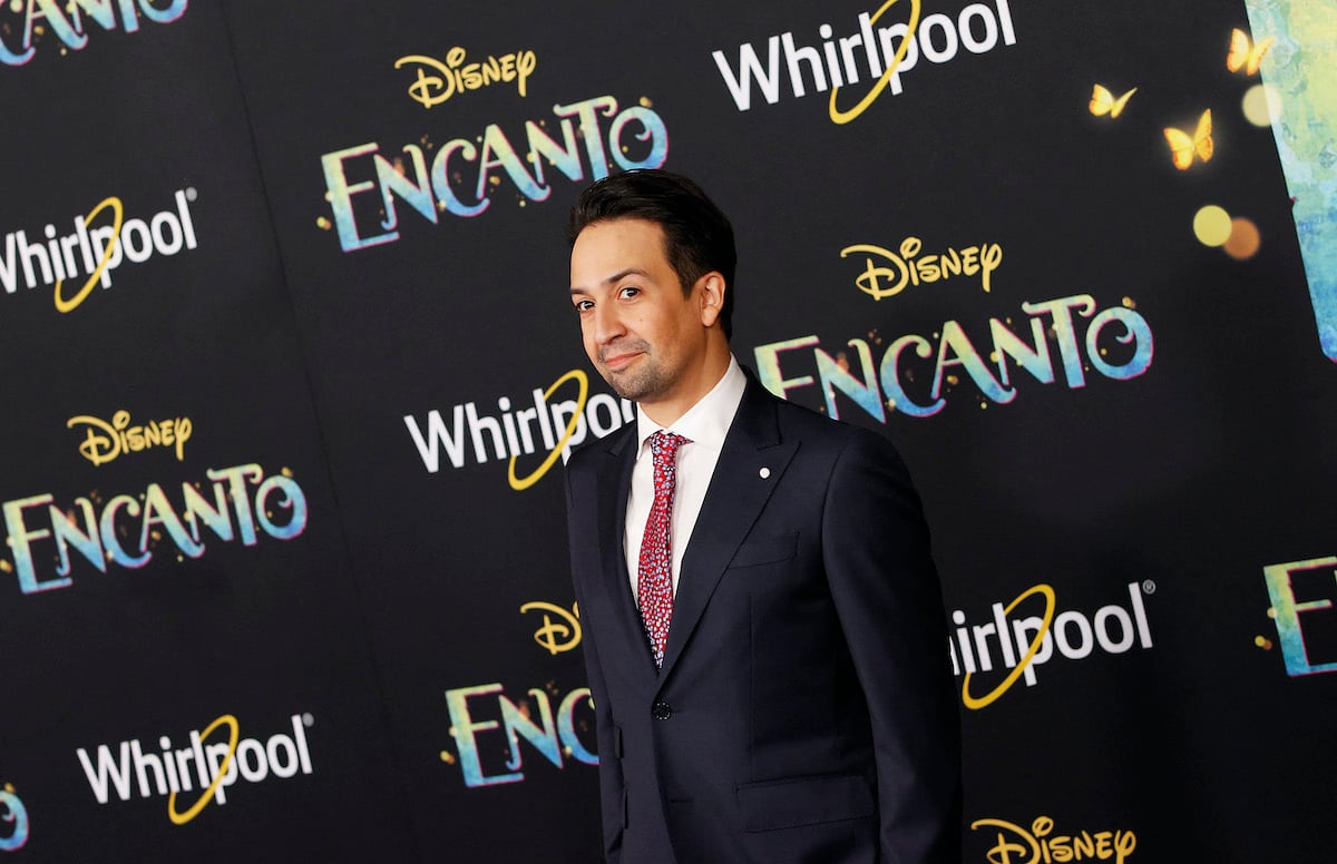 Lin-Manuel Miranda poses in a dark suit in front of the ‘Encanto’ logo