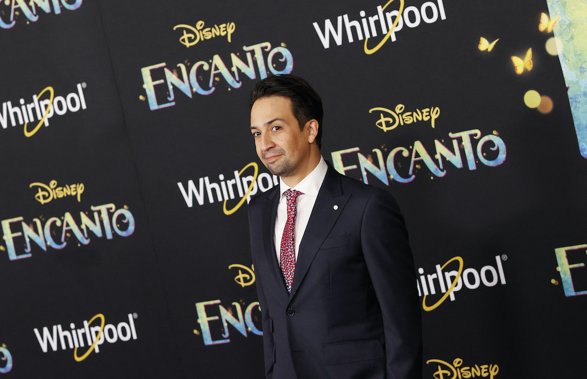 Lin-Manuel Miranda poses in a dark suit in front of the ‘Encanto’ logo