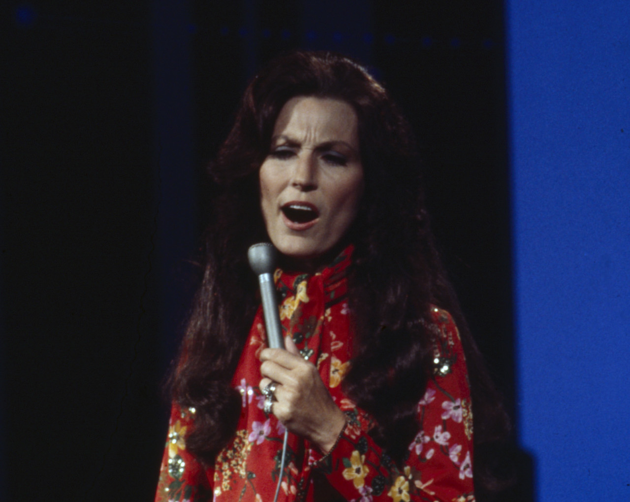Loretta Lynn singing into a microphone, wearing a red floral dress