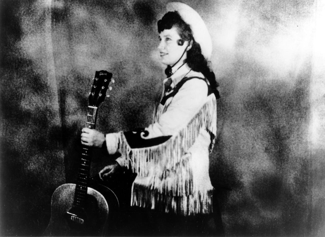 Loretta Lynn wears a cowboy hat and a fringe western style jacket while holding an acoustic guitar as she poses for a portrait, c. 1960
