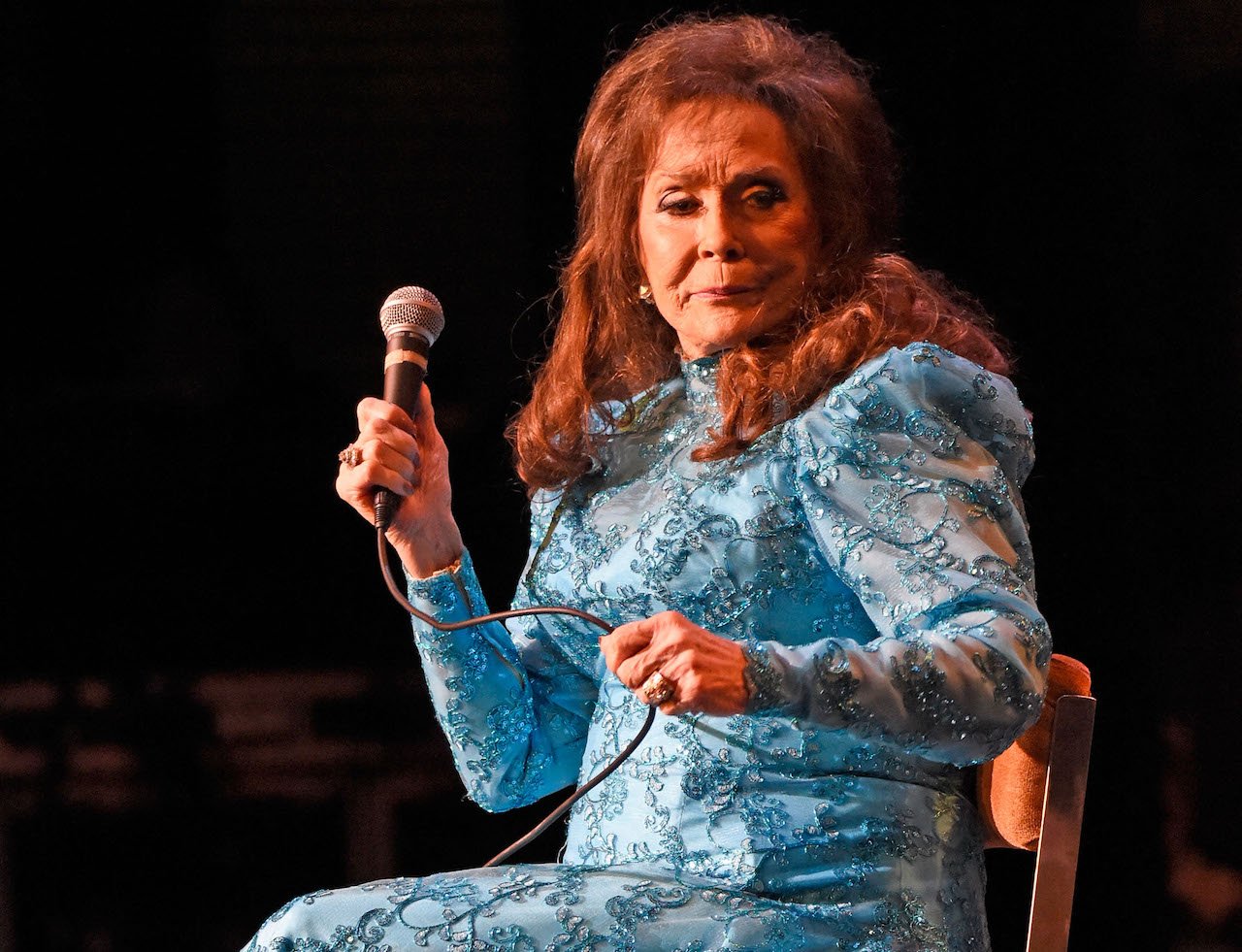 Loretta Lynn sits in a sparkly blue ball gown, holding a microphone in one hand and clenching the cord in her other fist
