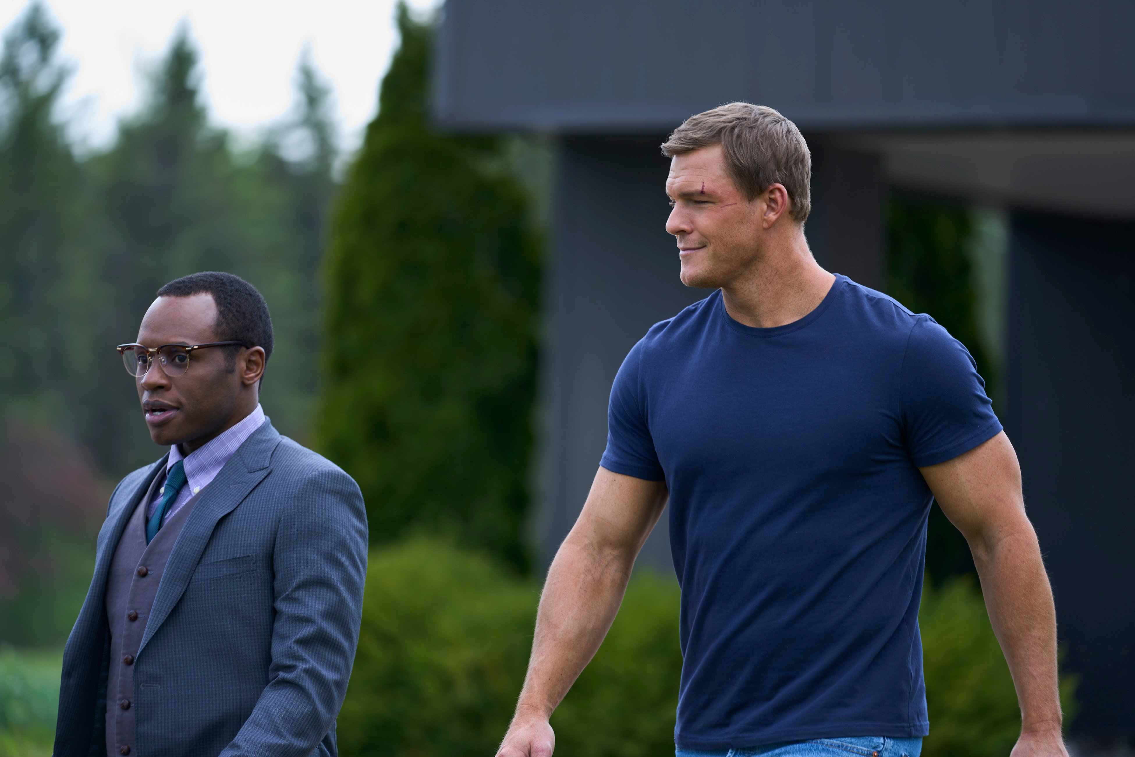Malcolm Goodwin, wearing a suit, standing next to Alan Ritchson, wearing a blue T-shirt, in an episode of 'Reacher'