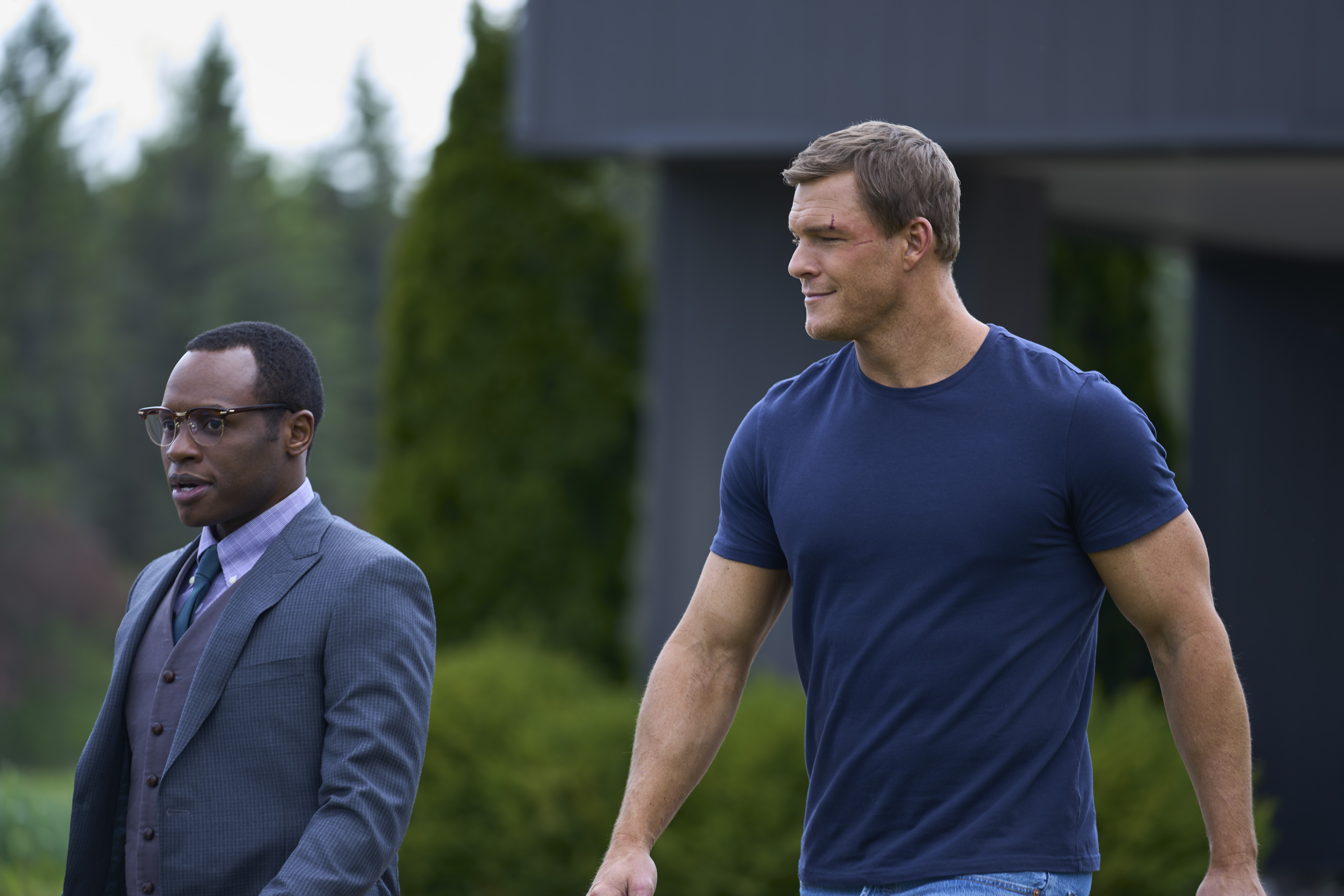 Malcolm Goodwin, wearing a suit, standing next to Alan Ritchson, wearing a blue T-shirt, in an episode of 'Reacher' 