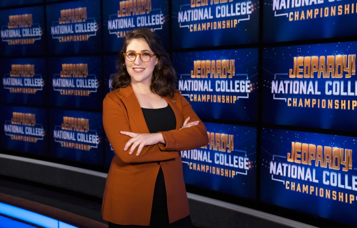 'Jeopardy!' host Mayim Bialik wears a brown jacket in front of the game show boards.