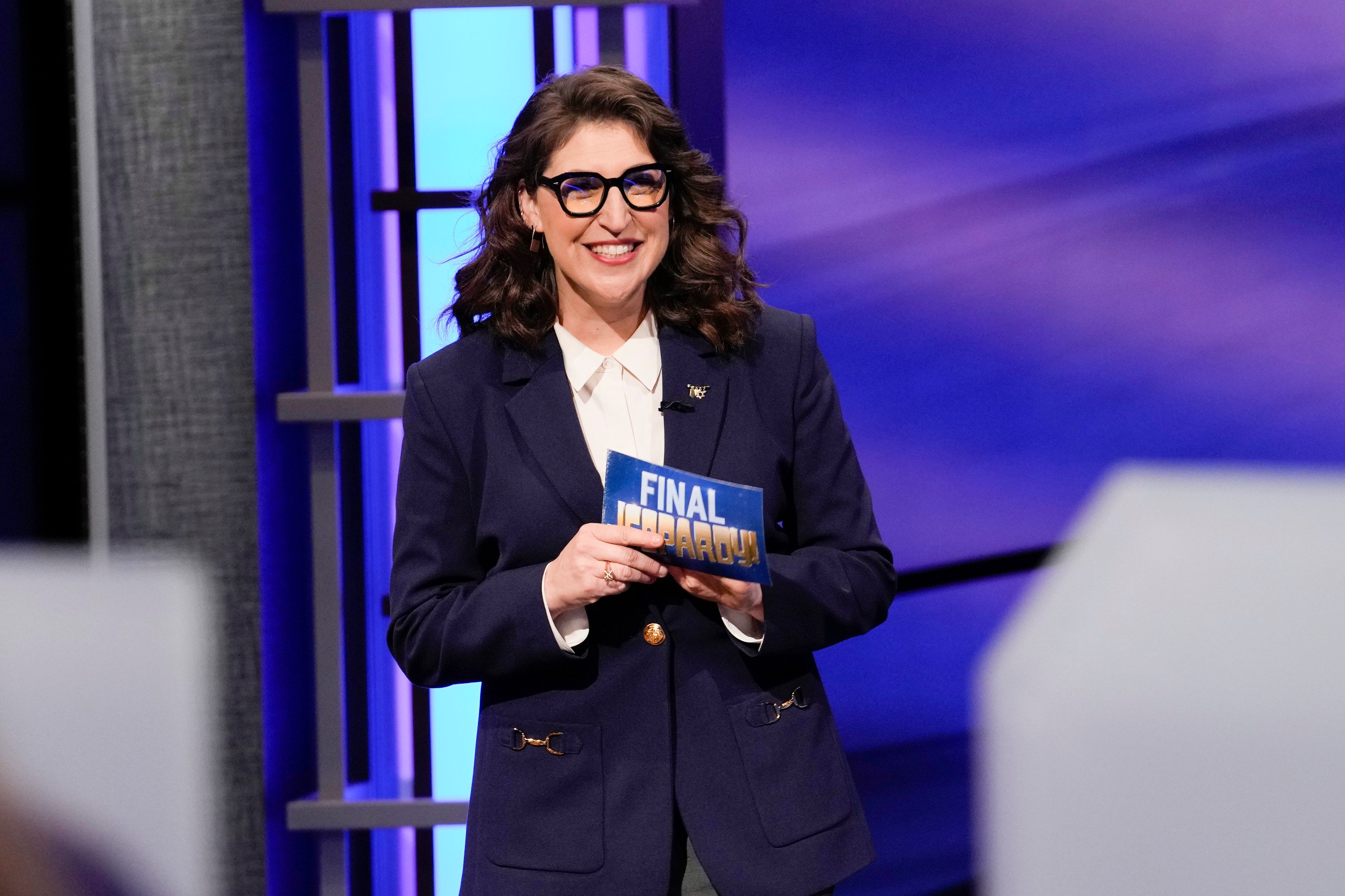 'Jeopardy! host Mayim Bialik holds a Final Jeopardy clue card in her hand on the game show's set.