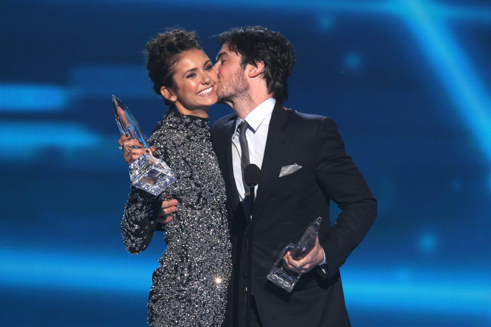Ian Somerhalder kisses his 'The Vampire Diaries' co-star, Nina Dobrev, on the cheek while accepting a People's Choice Award. Somerhalder wears a black suit over a white button-up shirt and gray tie. Dobrev wears a diamond studded long-sleeved dress.
