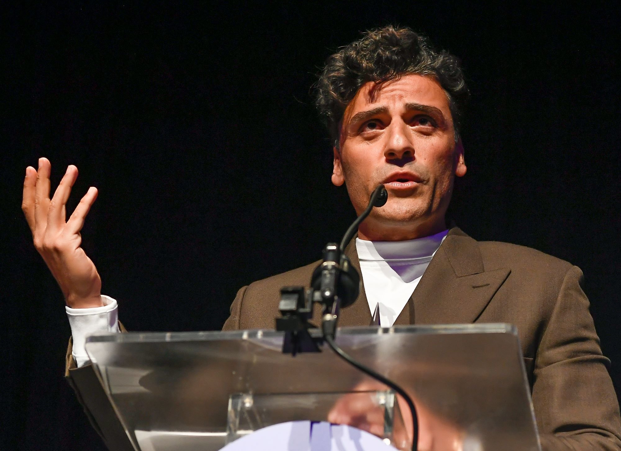 Oscar Isaac, who stars alongside Ethan Hawke in 'Moon Knight,' wears a brown suit over a white shirt while speaking at a podium.