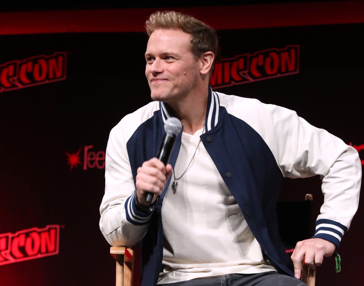 Outlander Sam Heughan wearing a jacket, white shirt and holding a microphone speaks onstage during Day 3 of New York Comic Con 2021 at Jacob Javits Center on October 09, 2021