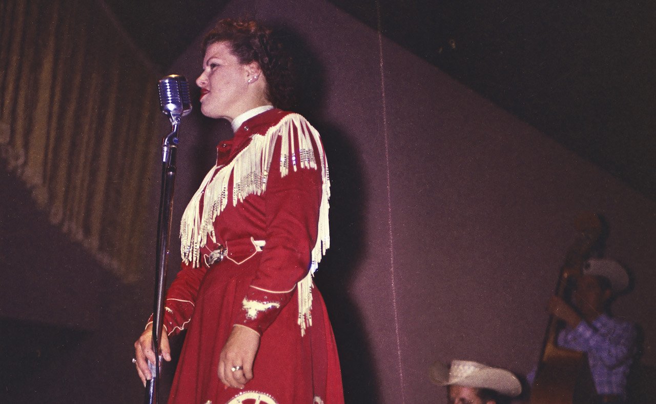 Patsy Cline sings in front of a microphone, wearing a red dress with white fringe
