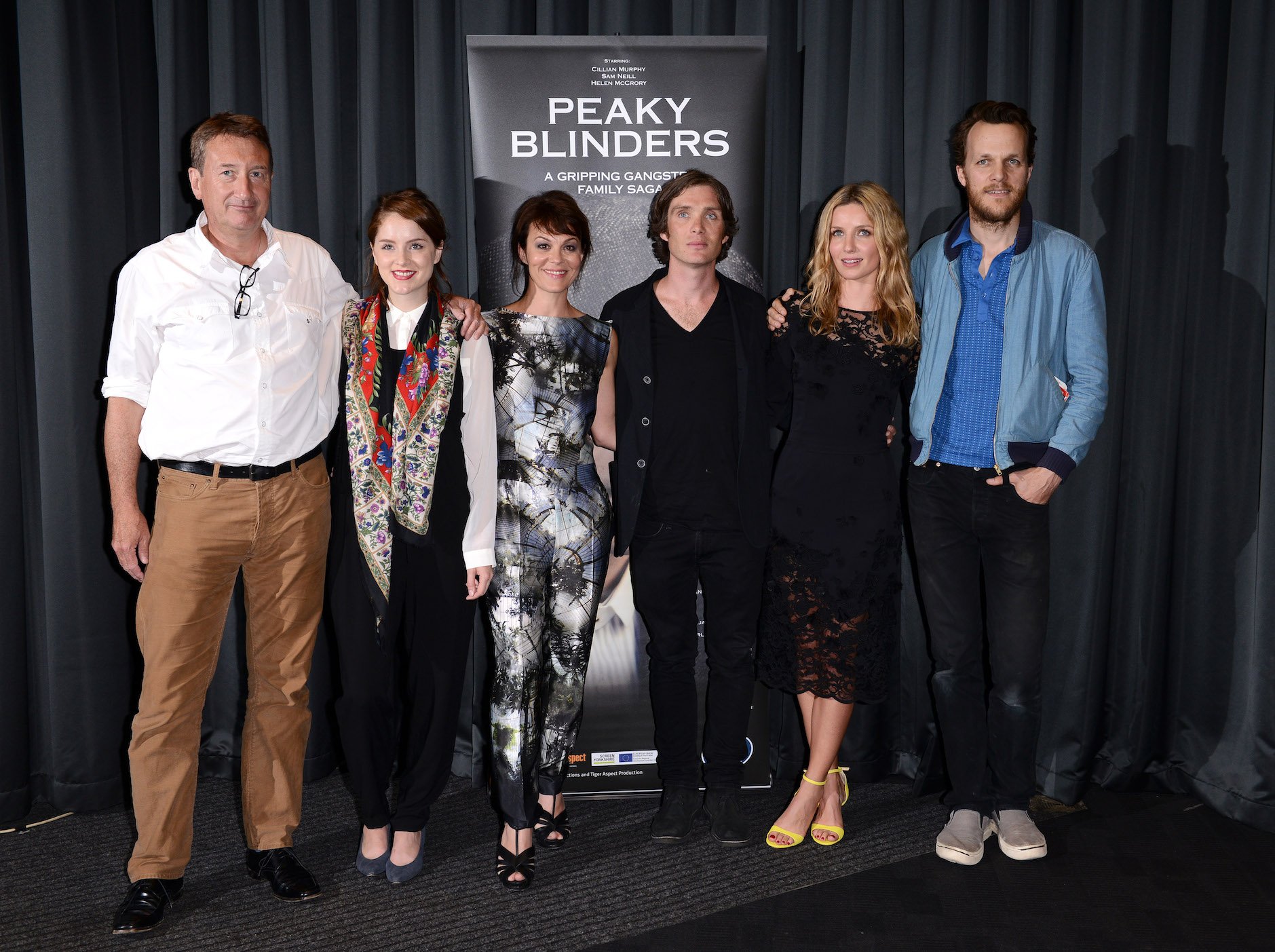 Steven Knight with some members of the 'Peaky Blinders' Season 6 cast, including Sophie Rundle, Helen McCrory, Cillian Murphy, Annabelle Wallis, and director Otto Bathurst. They're standing with their arms around each other in front of a 'Peaky Blinders' poster