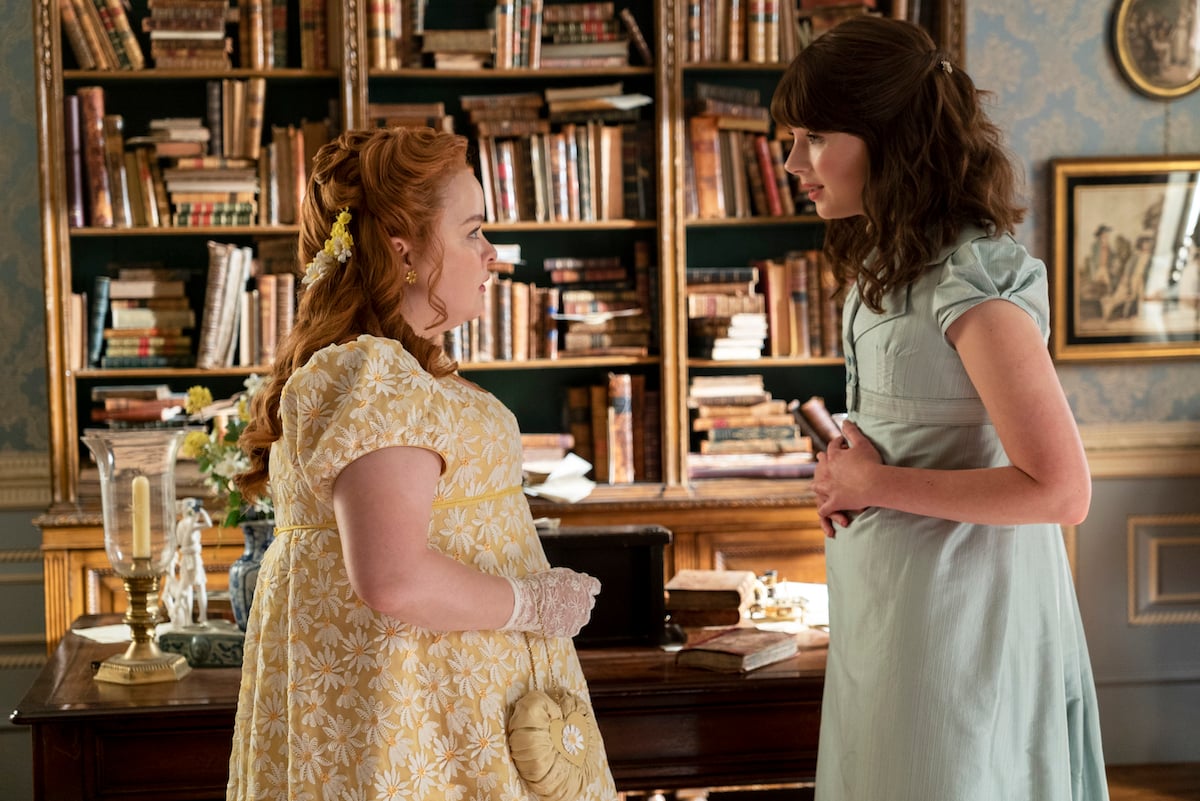Penelope, wearing a yellow dress, and Eloise, wearing a light blue dress, facing each other in an episode of 'Bridgerton' Season 2.