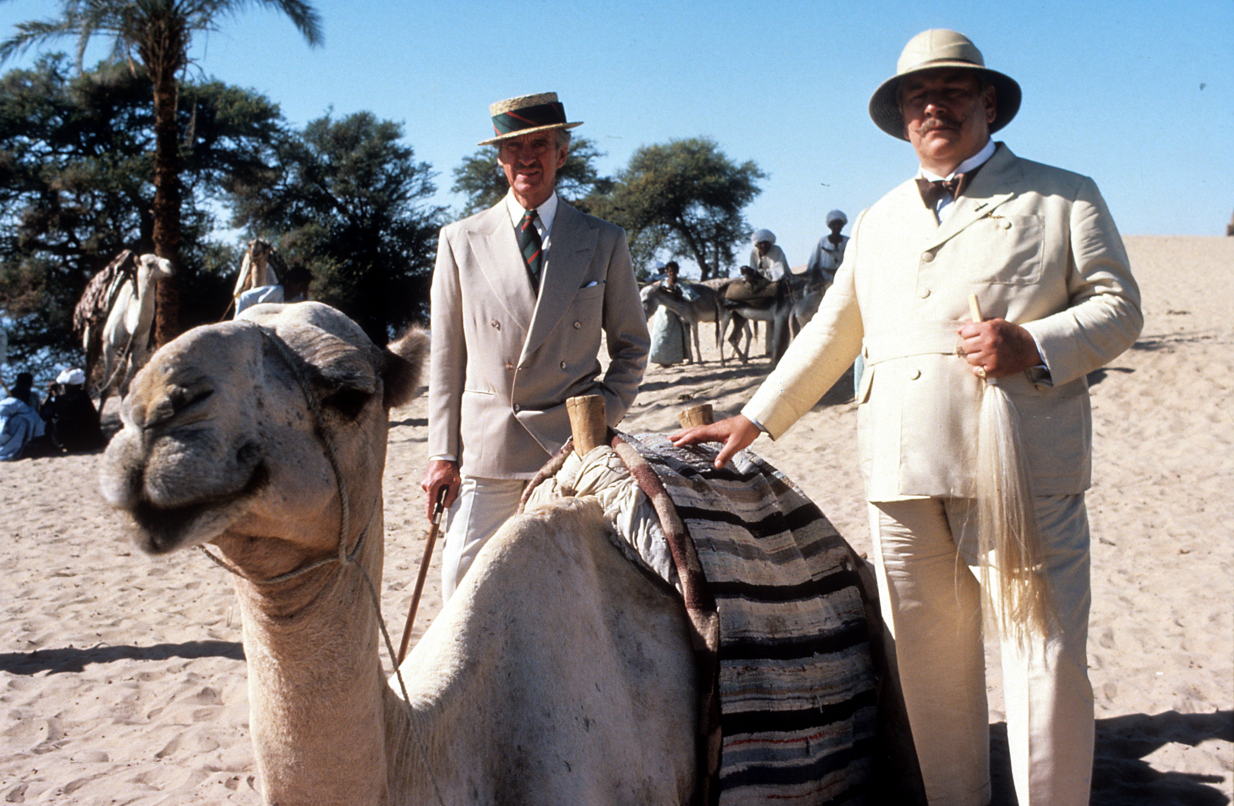 Peter Ustinov dressed in a suit and hat touching a camel, next to David Niven, in a scene from the film 'Death On The Nile', 1978