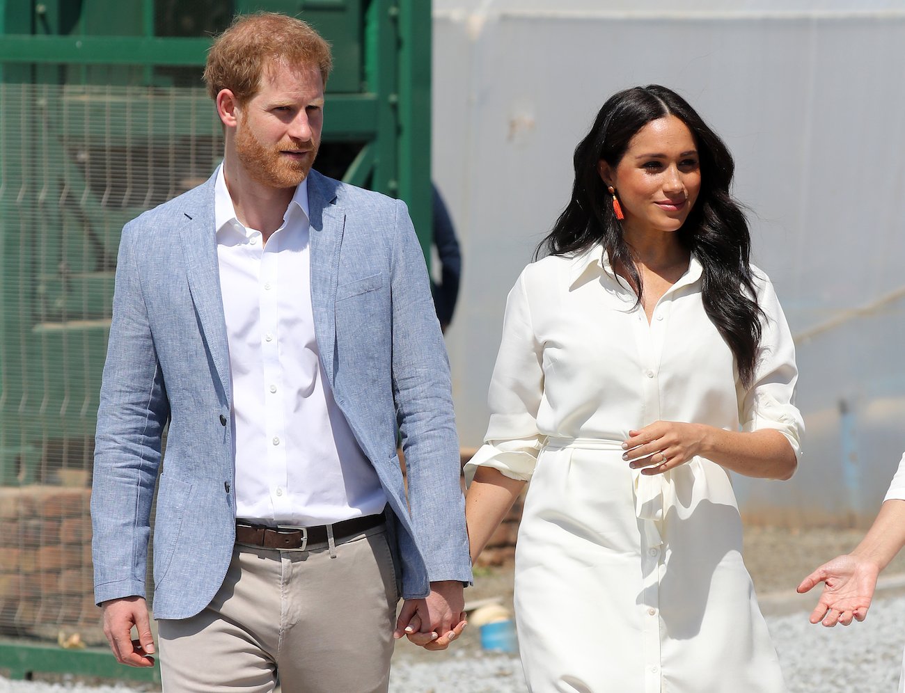 Prince Harry and Meghan Markle walking together holding hands with Harry wearing a light blue jacket and Meghan in a white shirt dress