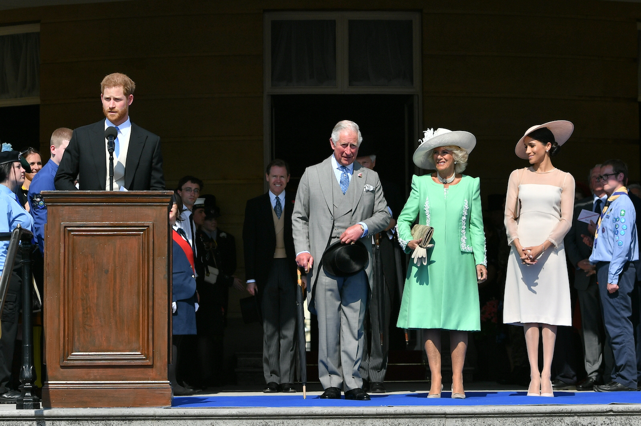 Prince Harry speaks behind a lectern while Prince Charles, Camilla Parker Bowles, and Meghan Markle look on