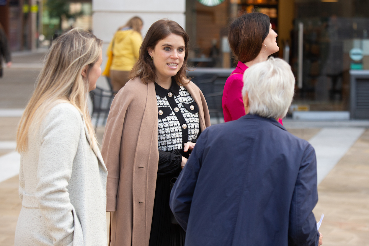 Princess Eugenie talks to bystanders wearing a black outfit and camel coat