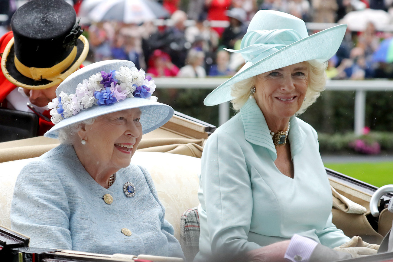 Queen Elizabeth and Camilla Parker Bowles wear pastel-colored outfits and hats riding in a carriage