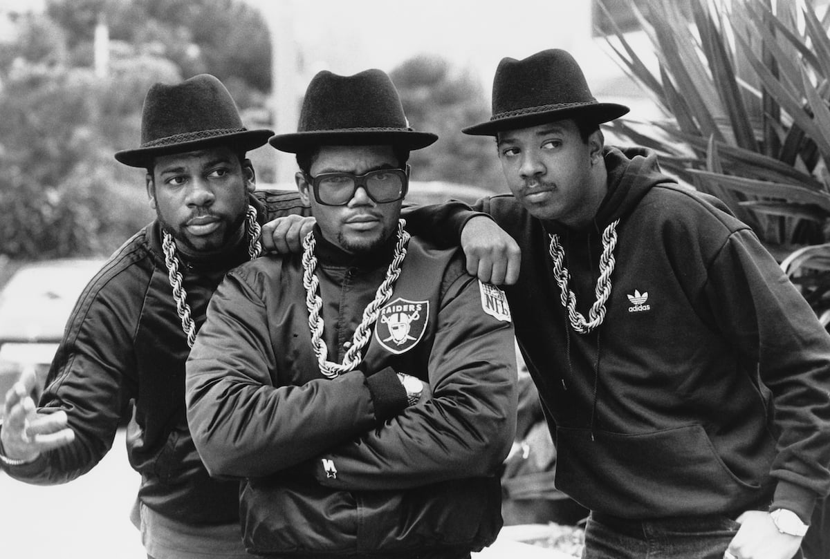 Run DMC members Jam Master Jay, DMC, and Rev Run wearing black hats and gold chains in a promotional photo.
