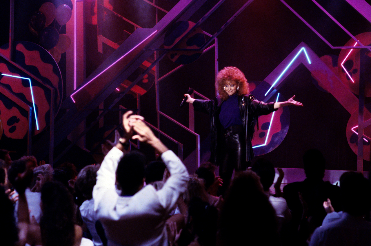 Reba McEntire performs on stage, holding her hands out to the side c. 1989