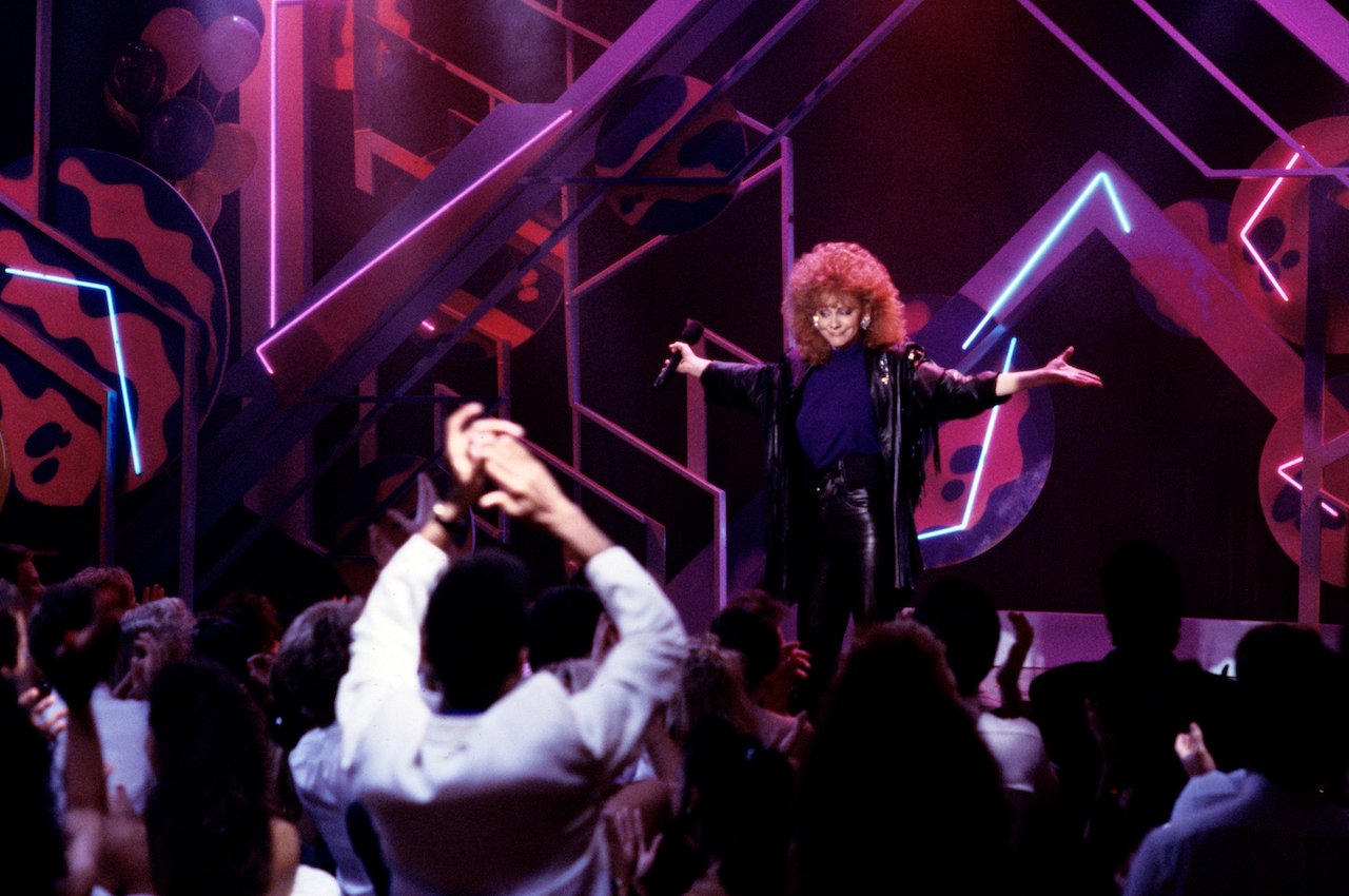 Reba McEntire performs on stage, holding her hands out to the side c. 1989