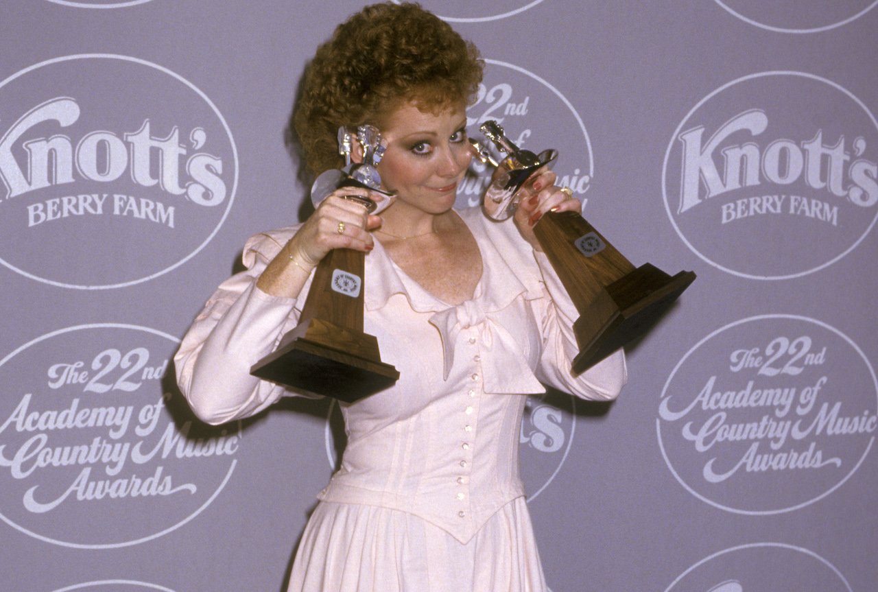 Reba McEntire in a light pink dress, holding two awards up beside her face and smiling