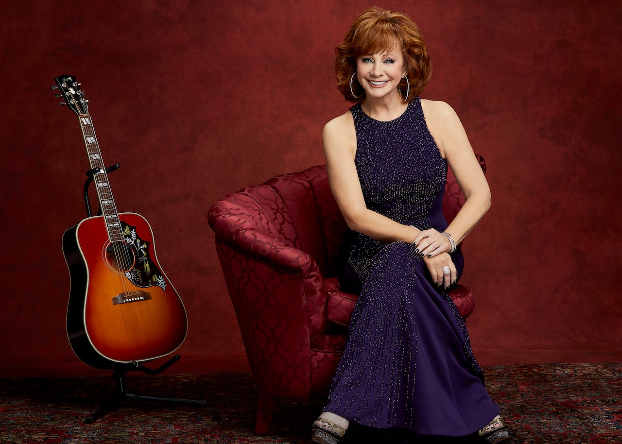 Reba McEntire in a dark blue dress, seated in a chair with her legs crossed next to a guitar