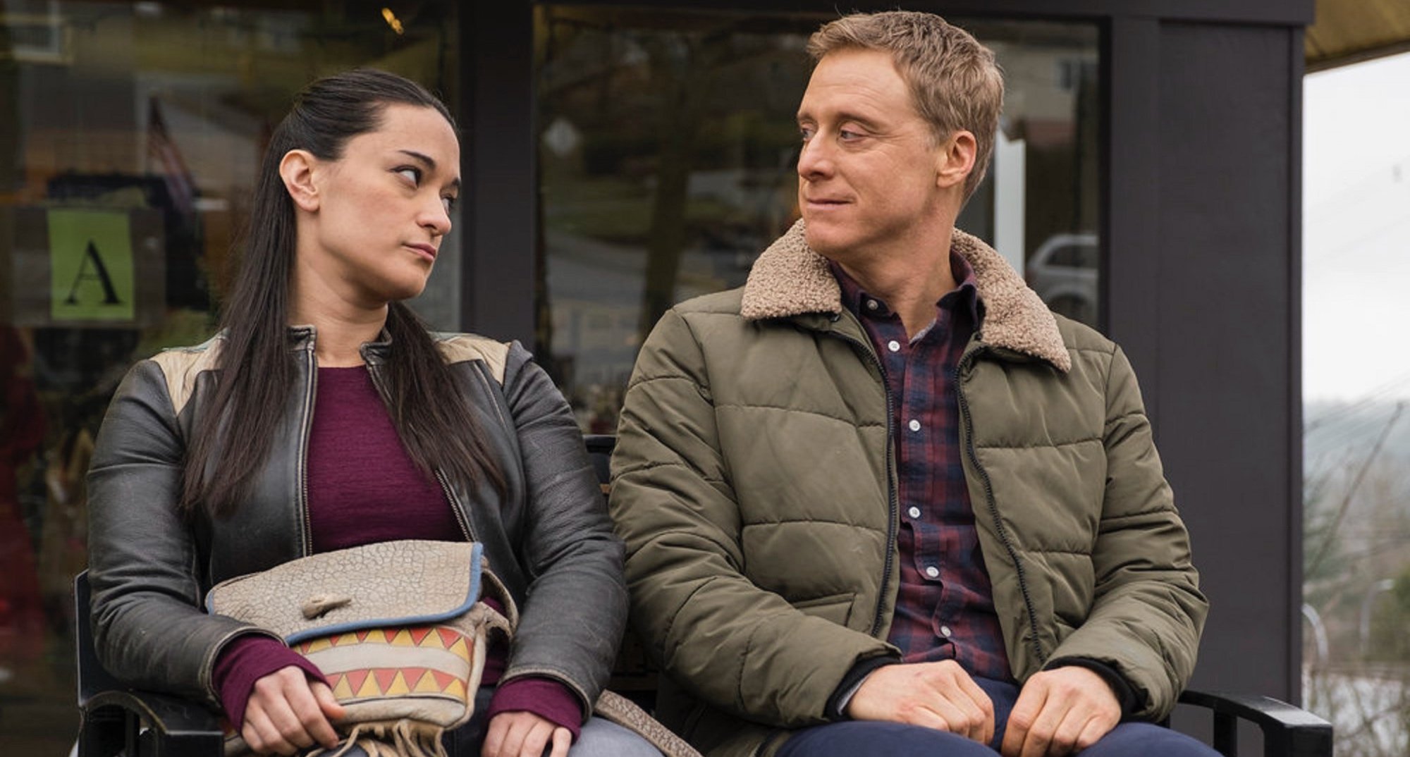 'Resident Alien' actors Sara Tomko and Alan Tudyk sitting on town bench.