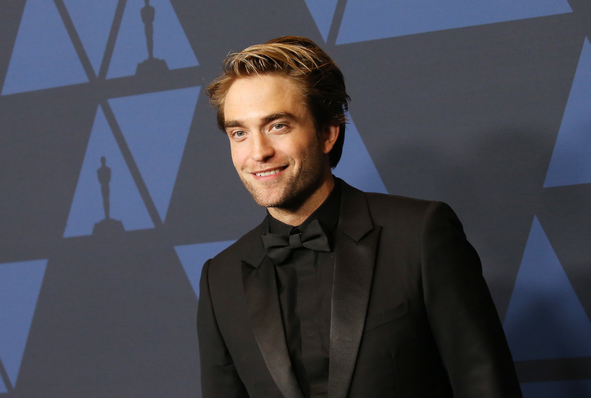 'The Batman' star Robert Pattinson. He's wearing a black suit and bowtie, standing in front of a black and blue wall, and smiling.