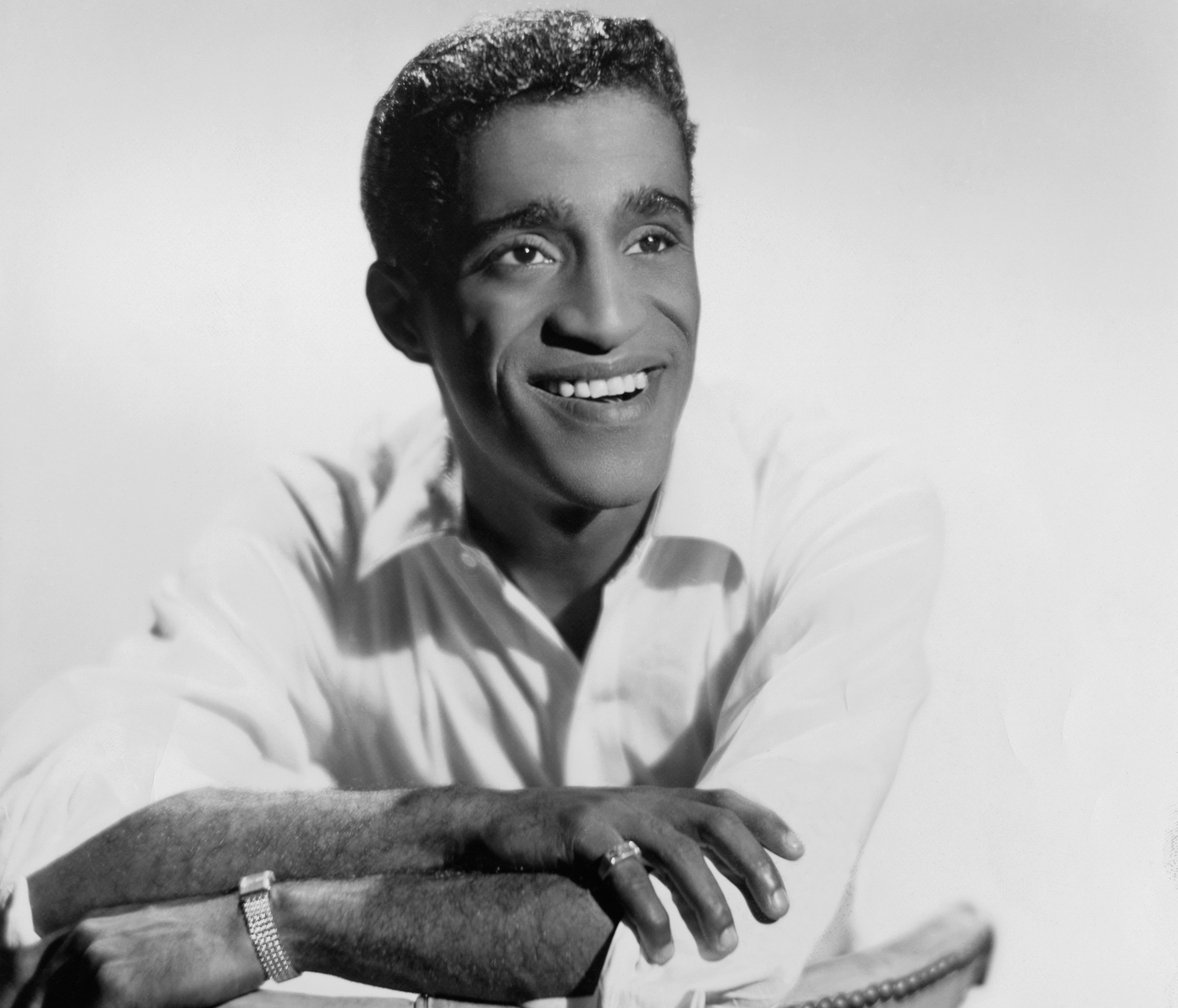Sammy Davis Jr. leans on a chair wearing a white shirt and watch.