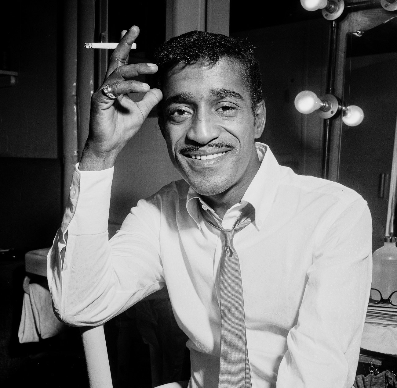 A black and white picture of Sammy Davis Jr. wearing a shirt and tie and holding a cigarette.