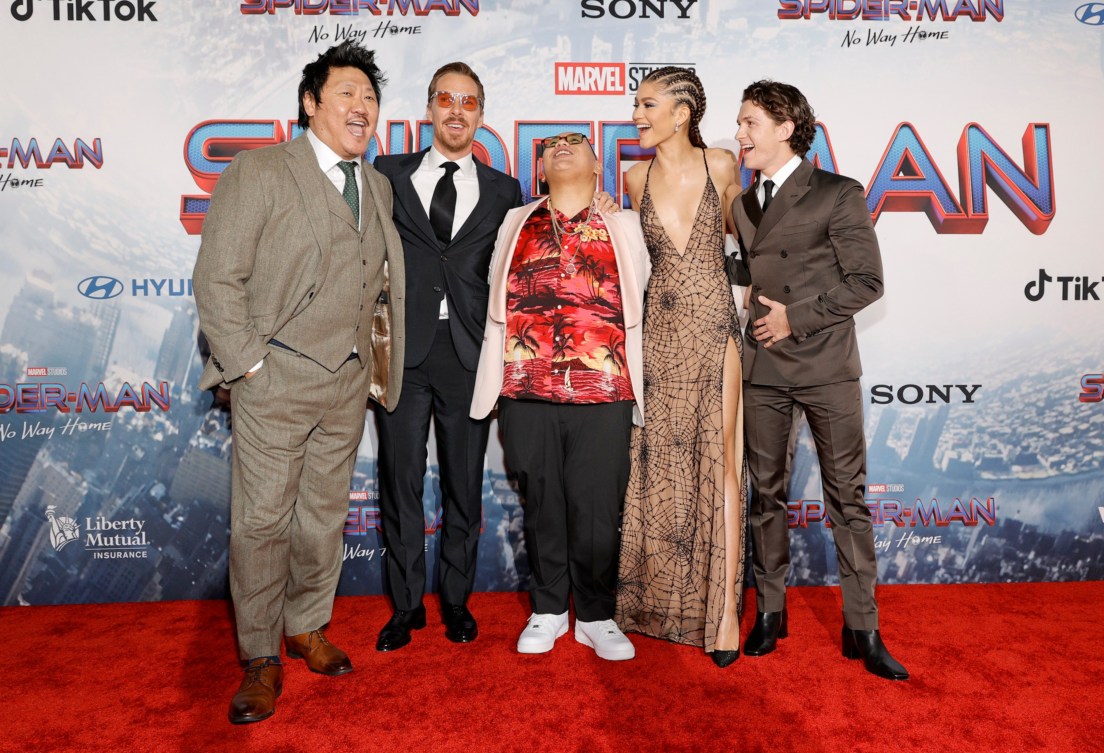 Benedict Wong, Benedict Cumberbatch, Jacob Batalon, Zendaya, and Tom Holland pose for pictures together on the red carpet for the premiere of 'Spider-Man: No Way Home,' which fans want to win the Fan-Favorite Award at the 2022 Oscars. Wong wears a gray suit. Cumberbatch wears a black suit. Batalon wears a white suit over a red Hawaiian shirt and black pants. Zendaya wears a tan dress with black spider webs. Holland wears a dark gray suit.