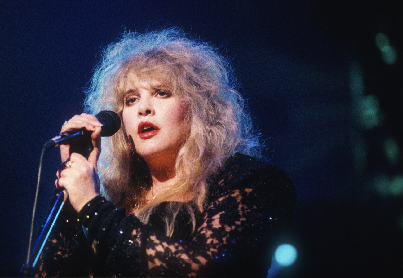 Stevie Nicks wears a black lace top and holds a microphone.