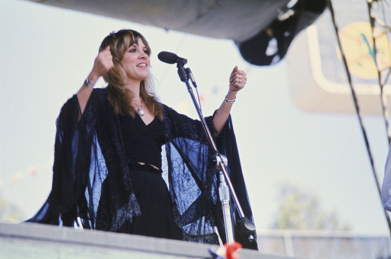 Stevie Nicks wears a black dress and shawl and stands in front of a microphone.
