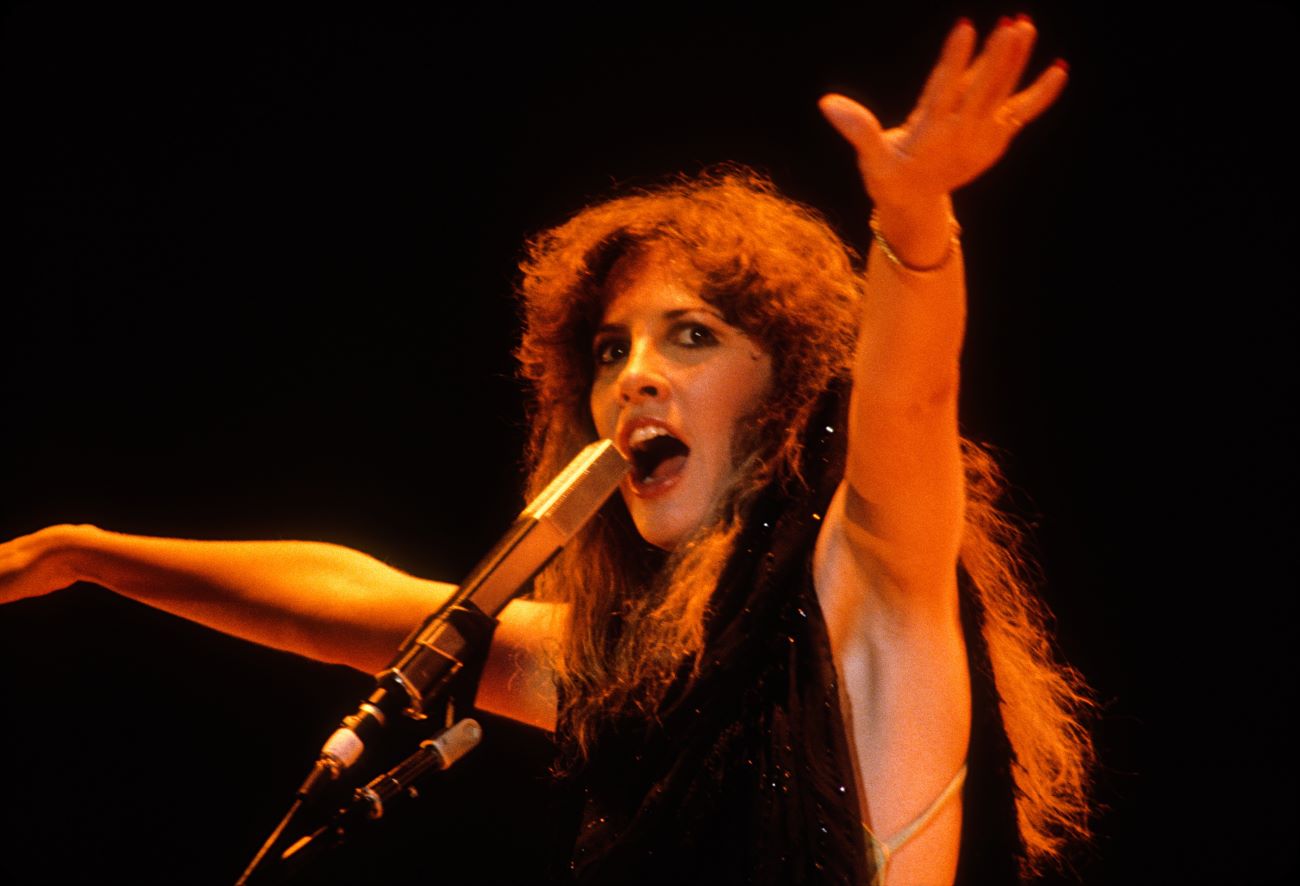 Stevie Nicks wears a black dress and sings a song into a microphone. She holds her arms in the air. 