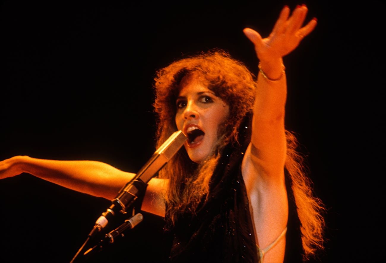 Stevie Nicks wears a black dress and sings a song into a microphone. She holds her arms in the air.