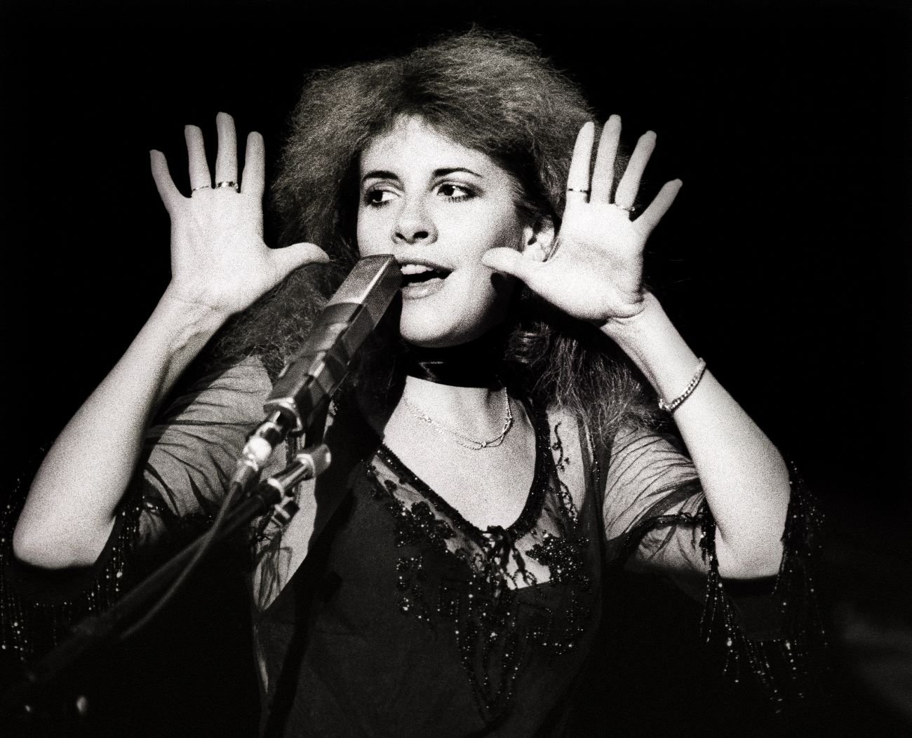 A black and white photo of Stevie Nicks wearing a lace top and standing in front of a microphone.