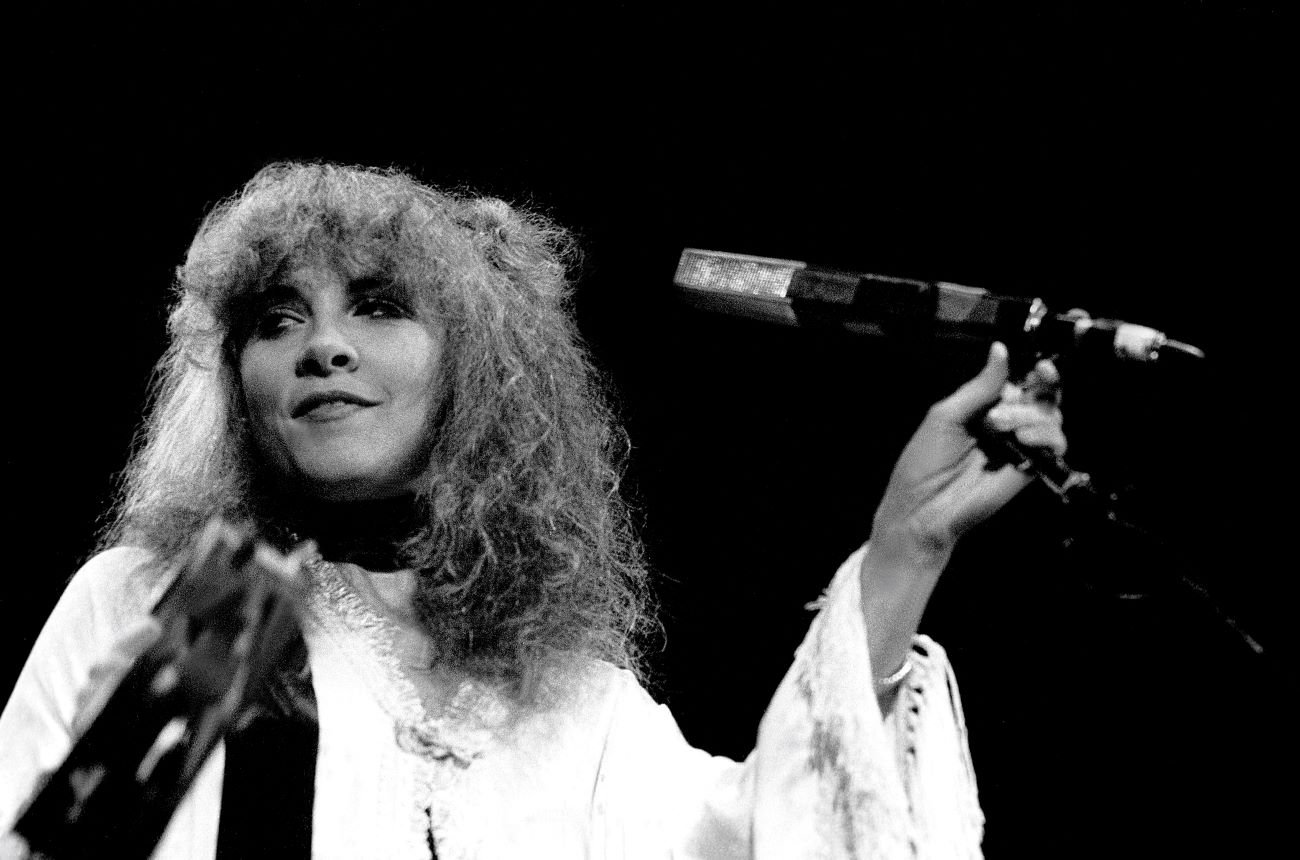 A black and white photo of Stevie Nicks wearing a white shirt and standing in front of a microphone.