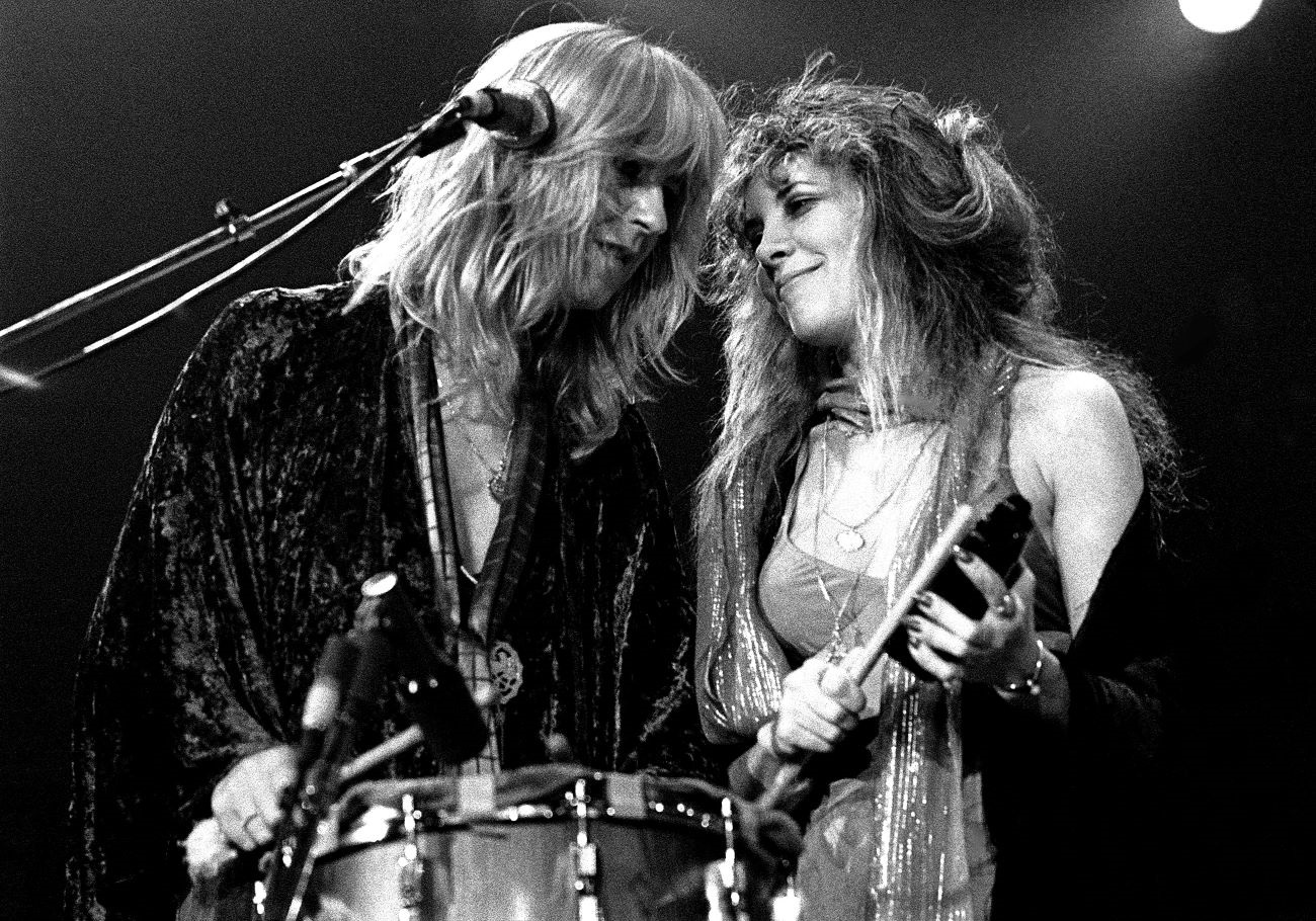 A black and white photo of Stevie Nicks and Christine McVie standing in front of a microphone with their heads close together.